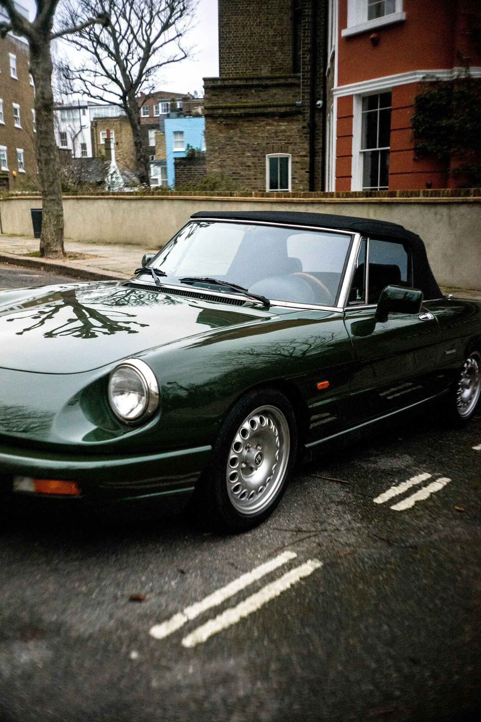 Green vintage convertible car parked on street with residential buildings and trees in the background.