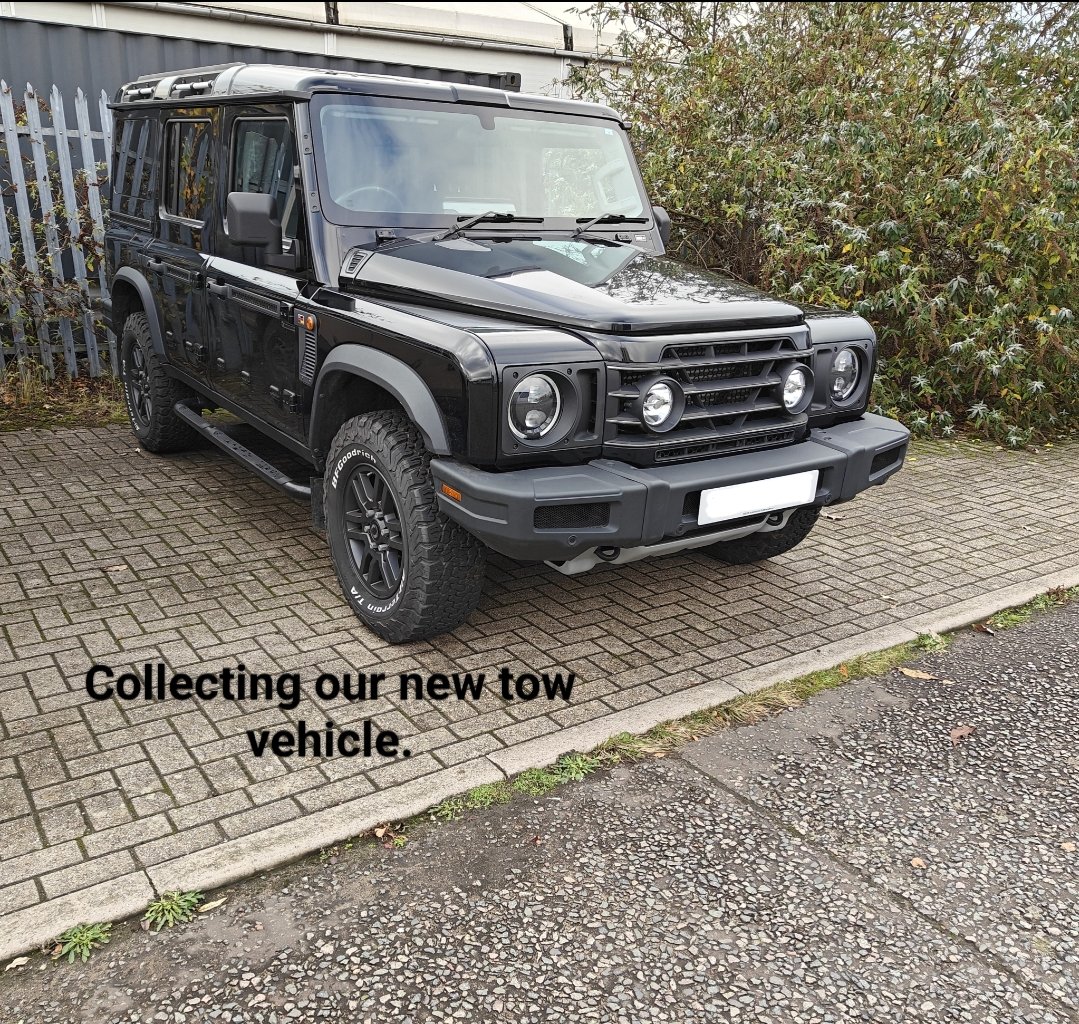 Black Land Rover Defender parked on a brick driveway next to bushes with a fence in the background. Text overlay says, "Collecting our new tow vehicle."