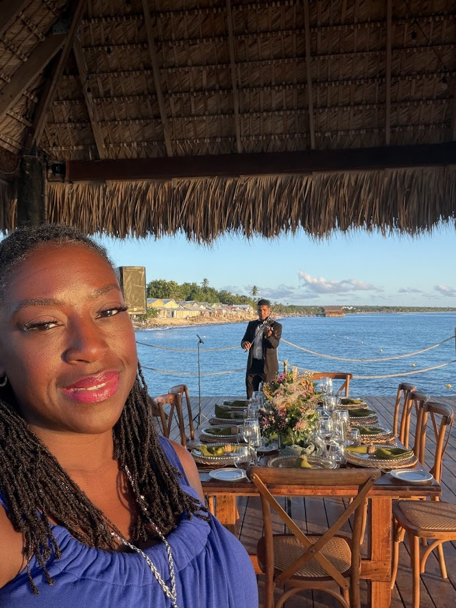 Black woman standing over a table setting overlooking the ocean with a violinist playing