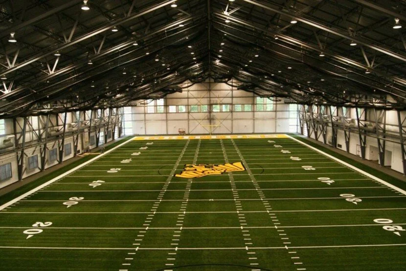 Indoor football field with green turf, white yard lines, and a center logo that says 'Arkansas.'