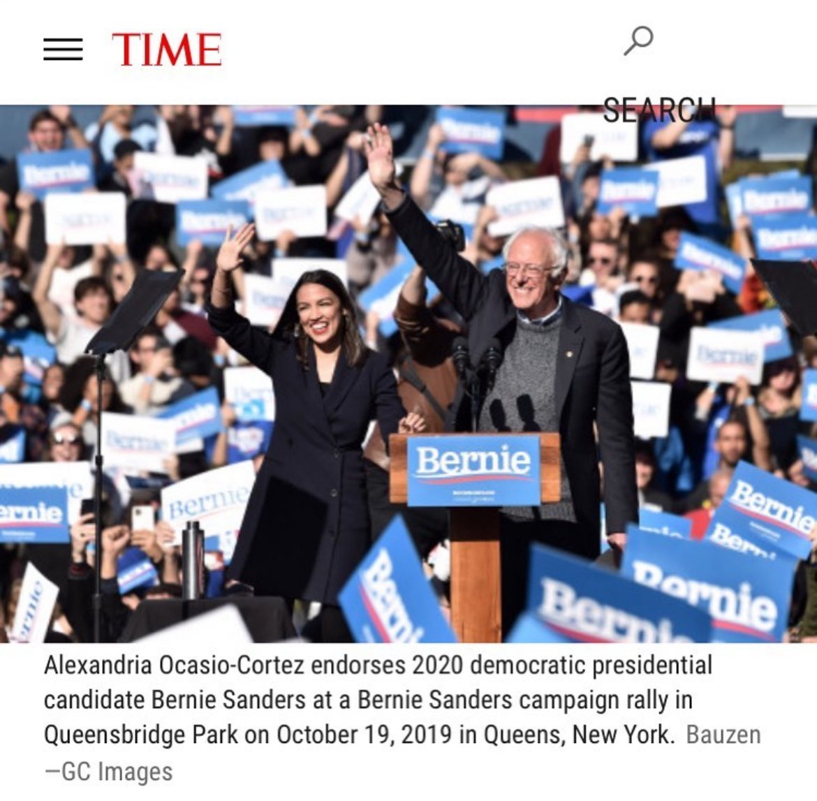 Alexandria Ocasio-Cortez and Bernie Sanders at a campaign rally, waving to the crowd, with many supporters holding Bernie Sanders signs in the background.