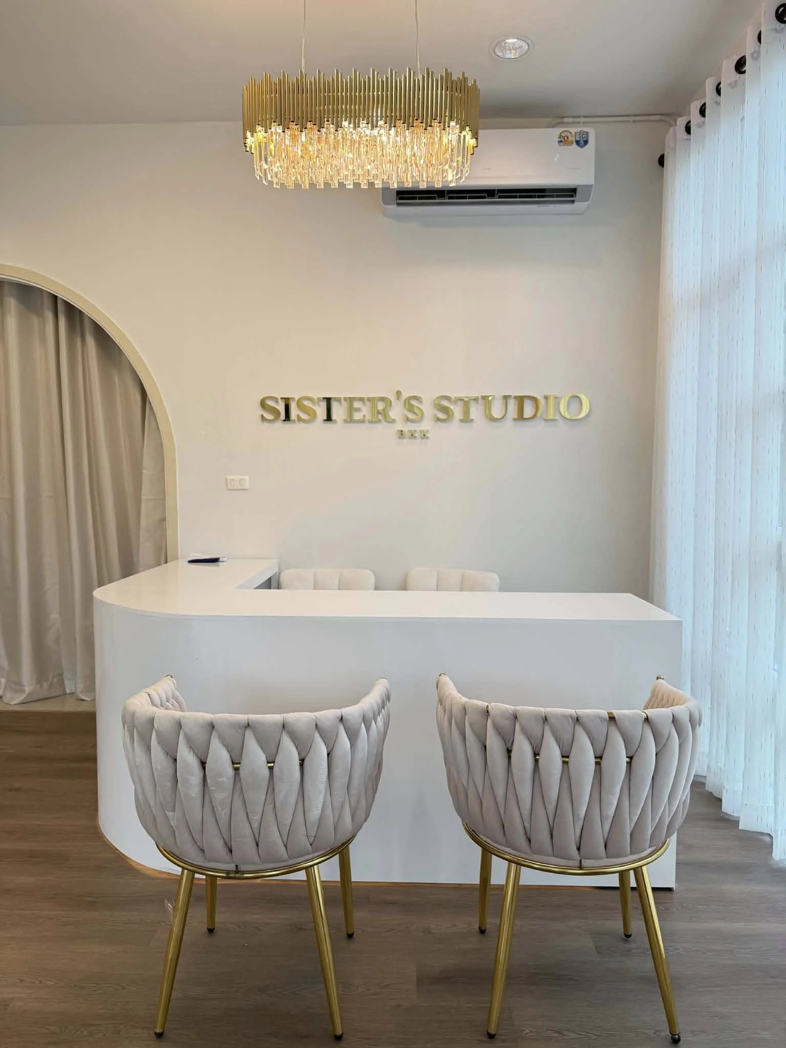 Interior of a beauty salon named Sister's Studio, showcasing a reception desk with three plush upholstered chairs and stylish decor, including a gold chandelier and white curtains.
