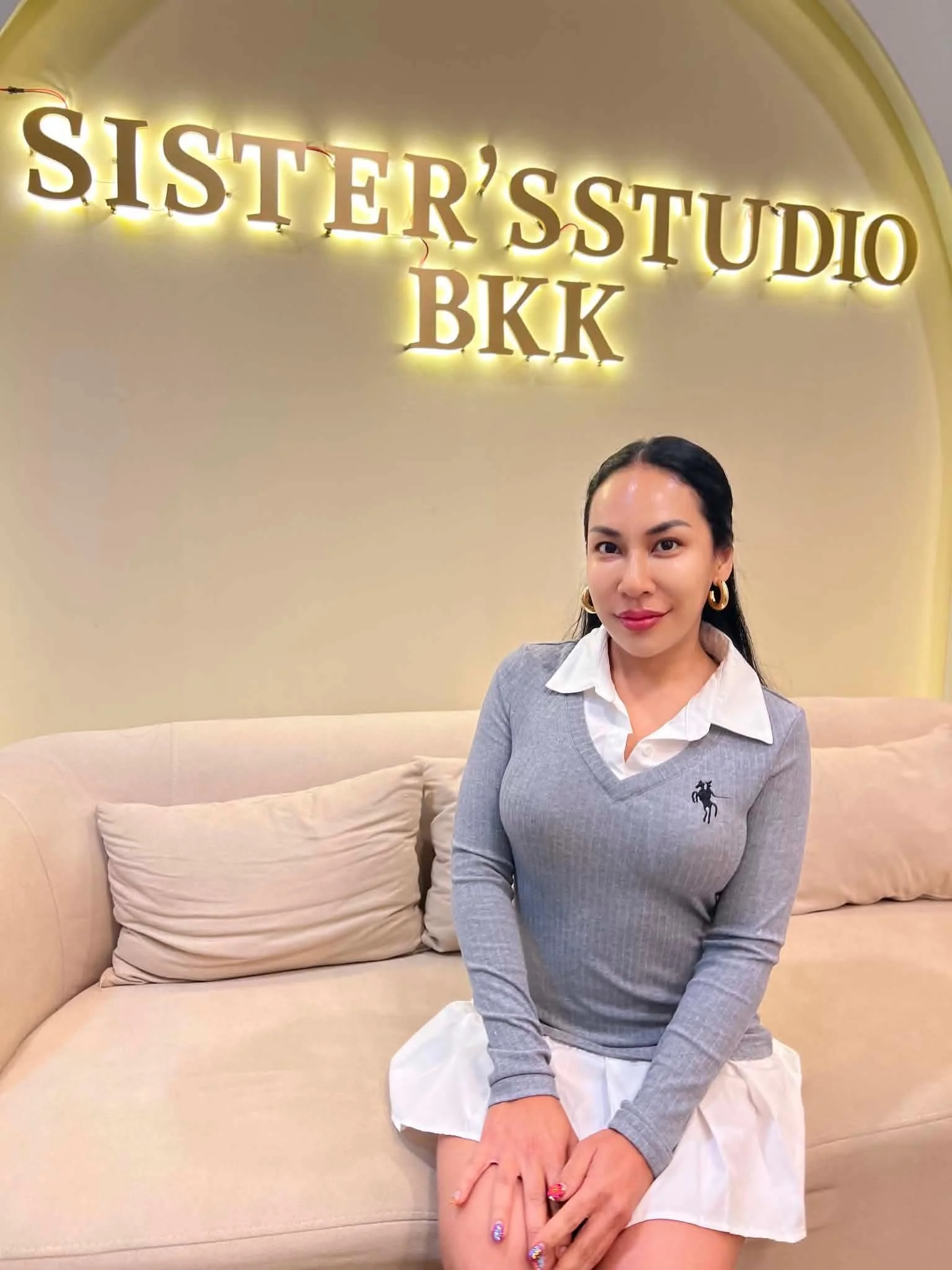 A woman with dark hair, wearing a light gray sweater with a black horse logo and a white skirt, sitting on a beige sofa in front of a yellow wall with a sign that reads 'SISTER'S STUDIO BKK' illuminated in yellow backlight.