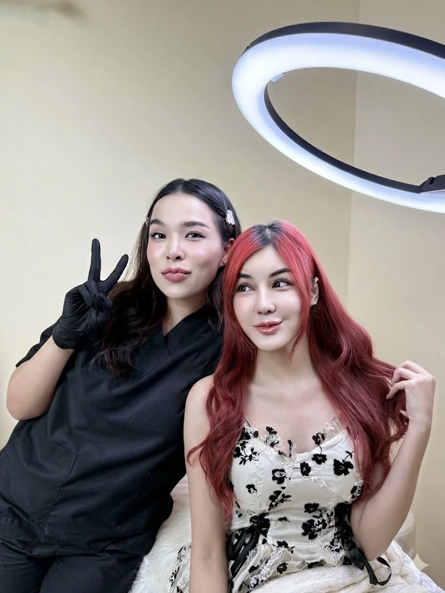 Two women sitting together indoors. One woman has dark hair, wearing black scrubs and a black glove, making a peace sign. The other woman has long red hair, wearing a white dress with black floral patterns, touching her hair.