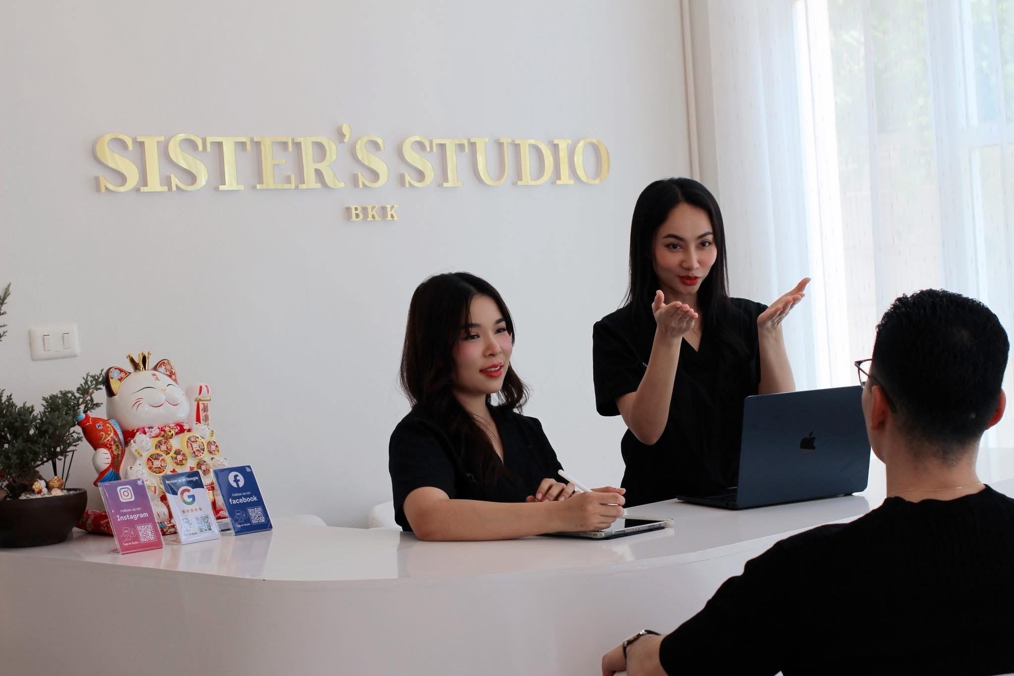Two women sitting at a white desk with one looking at a laptop and the other gesturing with her hands, in an office with a sign that reads 'SISTER'S STUDIO BKK' behind them. There are decorations and social media info cards on the desk.