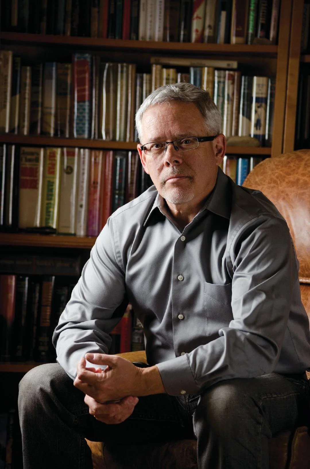 A middle-aged man with glasses and gray hair sitting on a brown leather chair in a room with a bookshelf filled with books in the background.