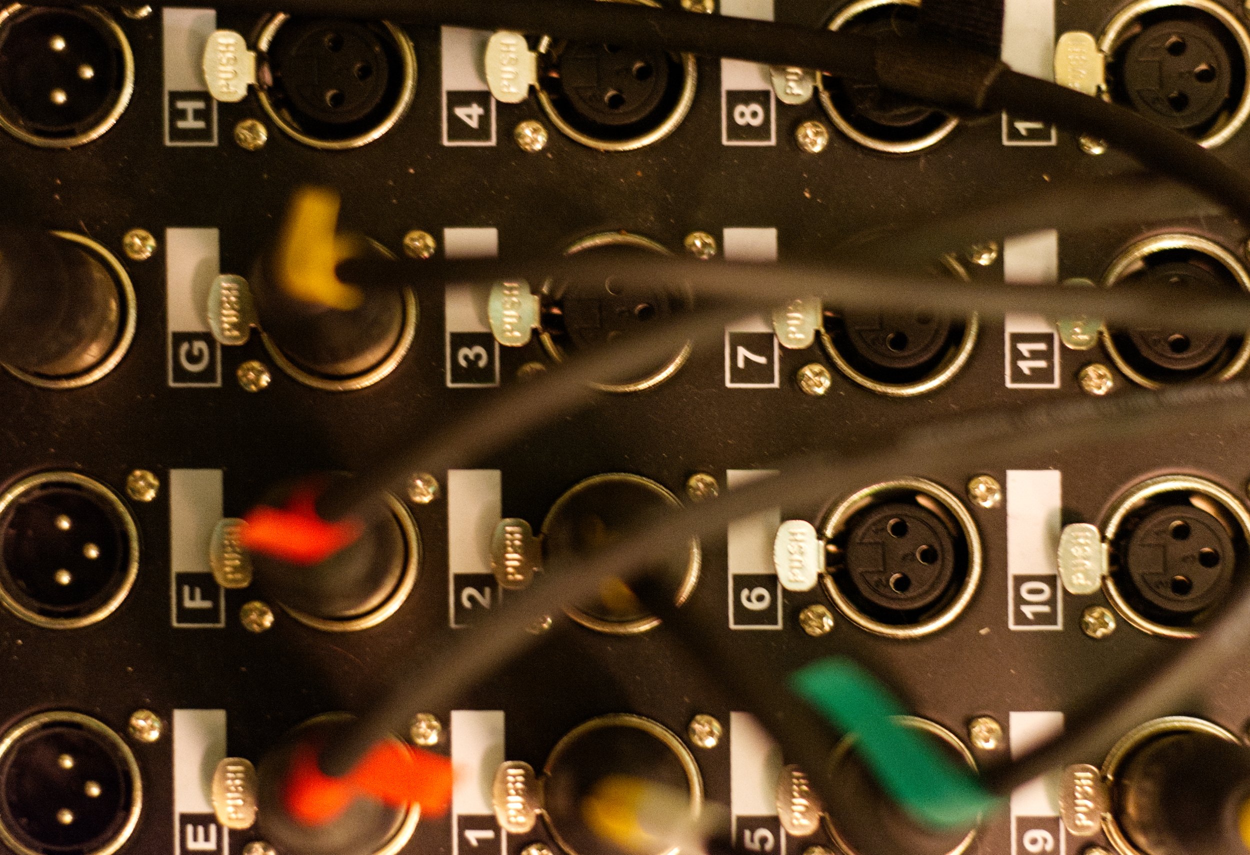 Close-up of a professional audio mixer with various input and output ports, some connected with colored cables, labeled with numbers and letters.
