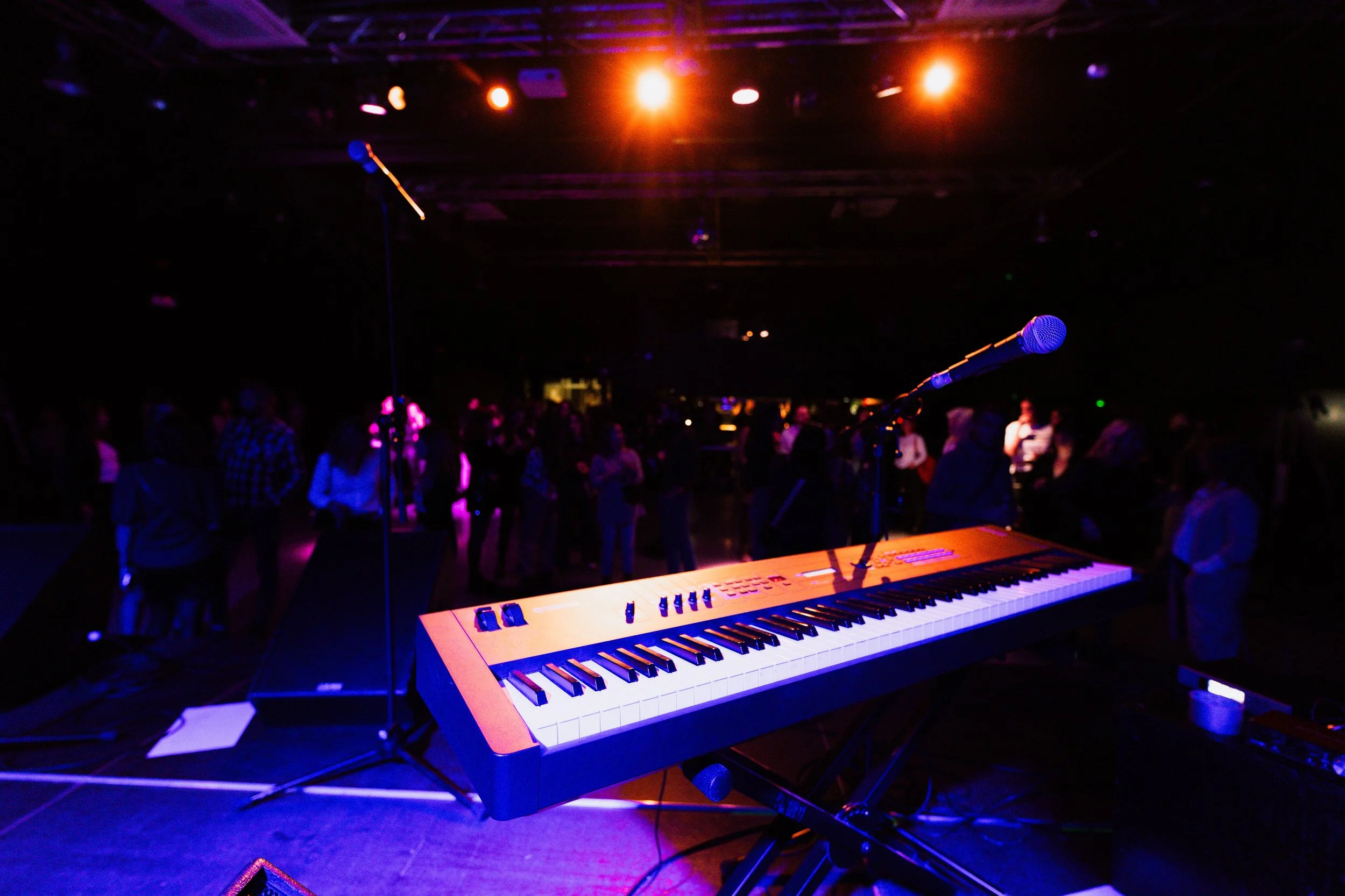 A digital keyboard piano on a stage with a microphone in front of it, under colorful stage lighting, and a crowd of people in the background at a live music event.