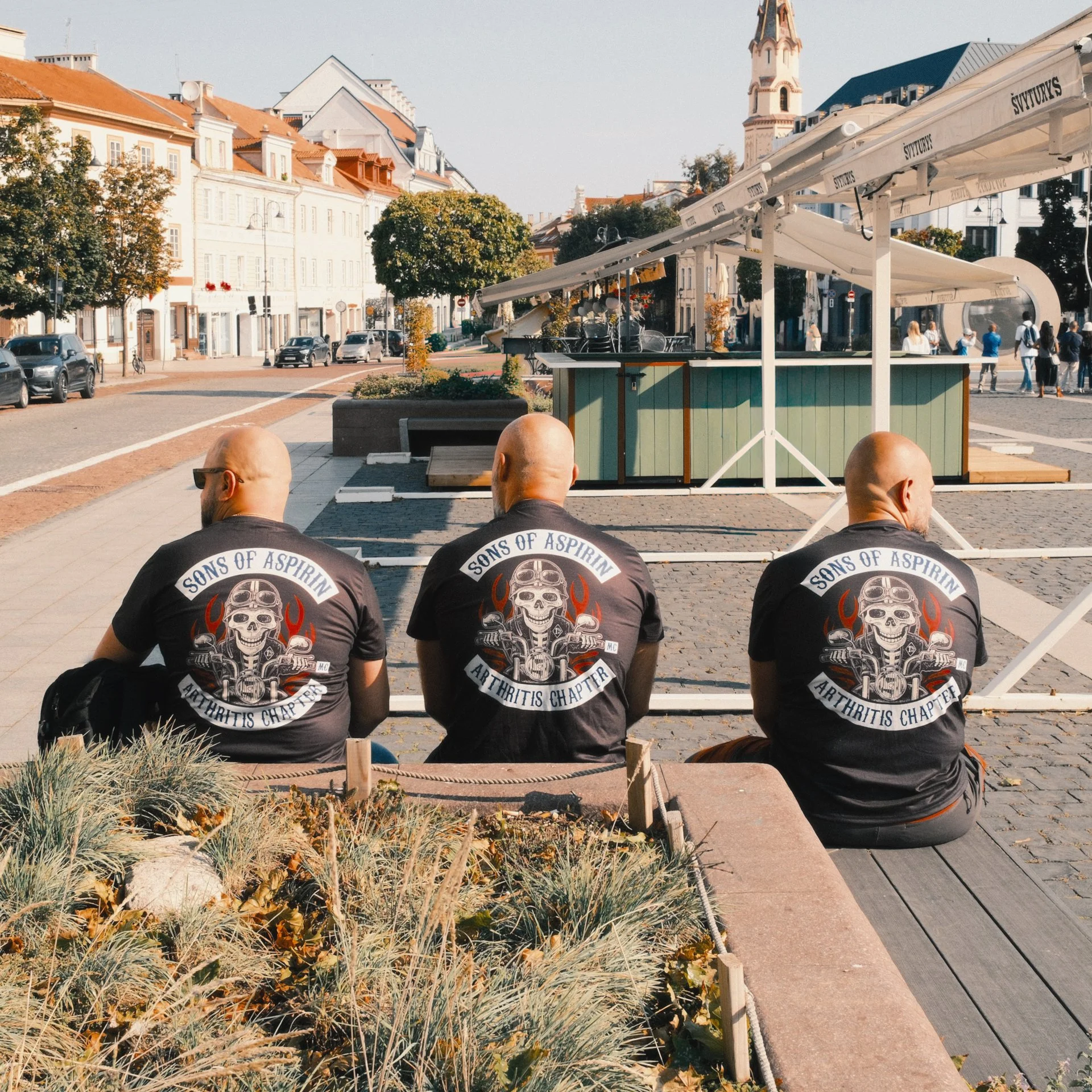 Three men with shaved heads sitting on a bench in a city square, wearing black T-shirts with a skull and flames graphic and the text 'Sons of Aspirin, Arthritis Chapter' on the back. A market stall is in the background, and there are buildings, trees