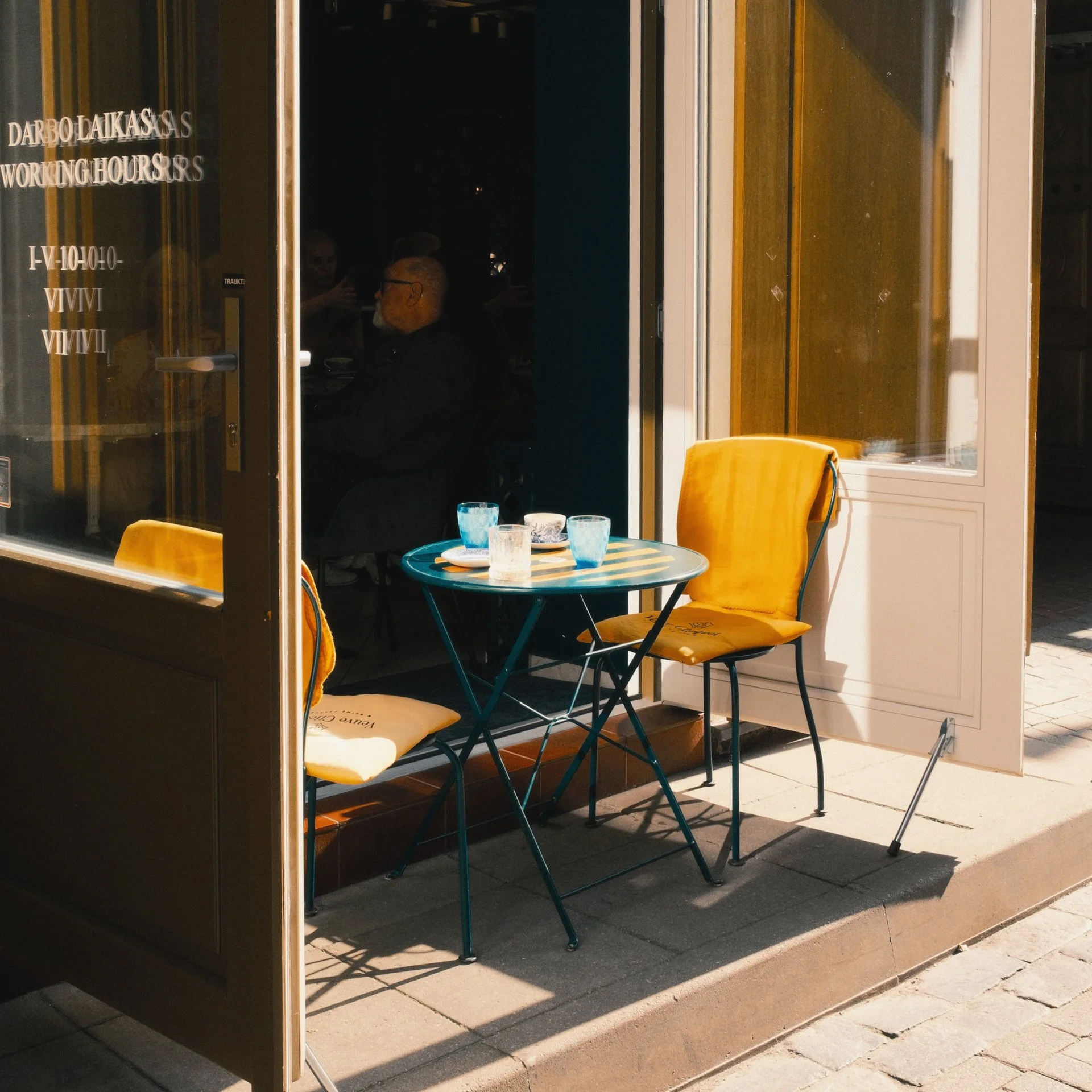 Empty outdoor cafe seating area with one table and two yellow chairs outside a cafe, with a person inside visible through the window.