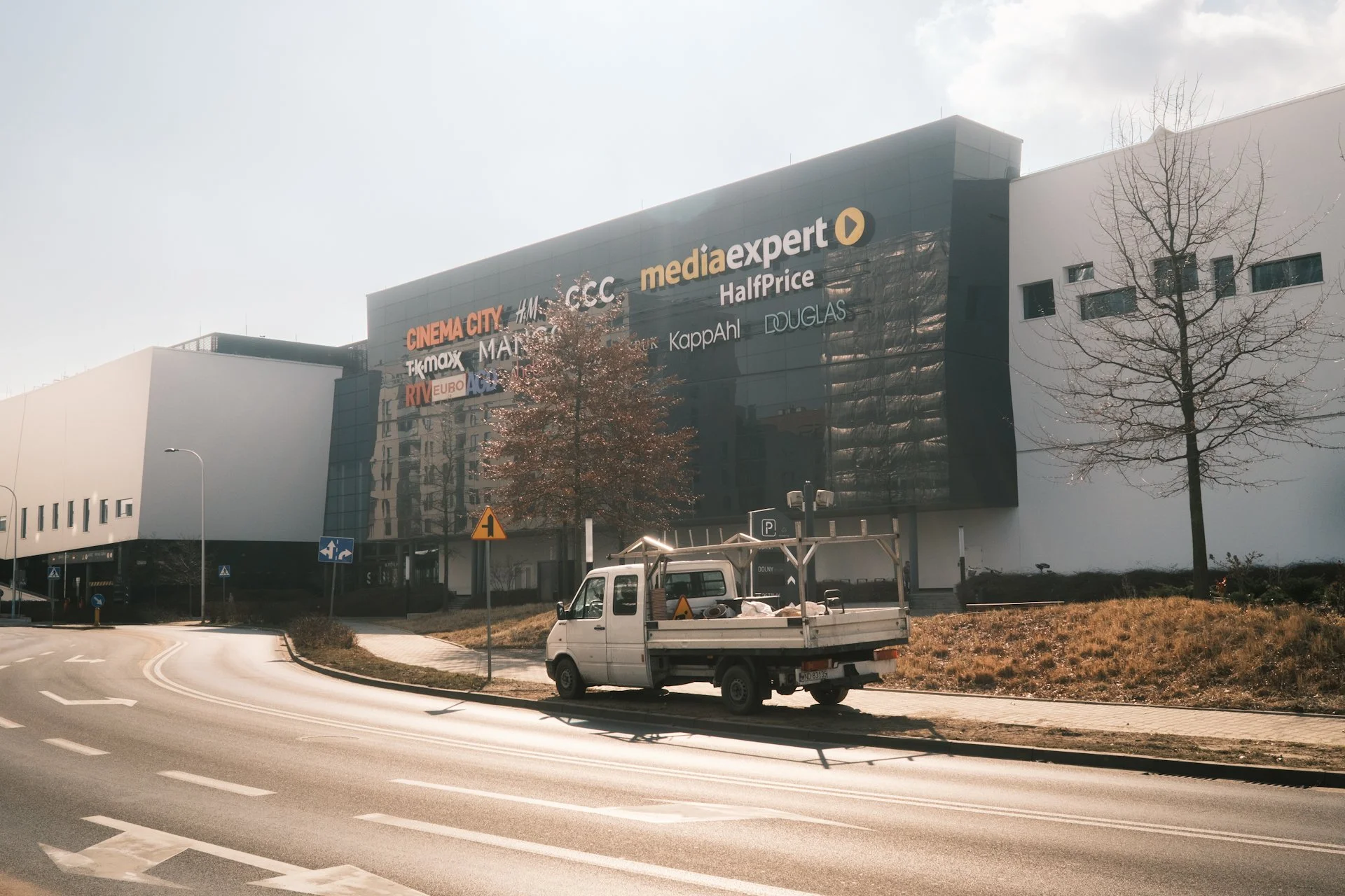 A commercial shopping mall with store signs, trees, and a parked small truck on the road.
