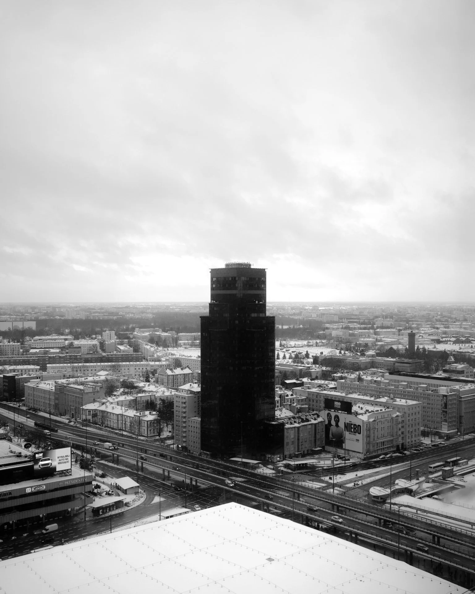 Black skyscraper surrounded by city buildings under cloudy sky, snow on rooftops.