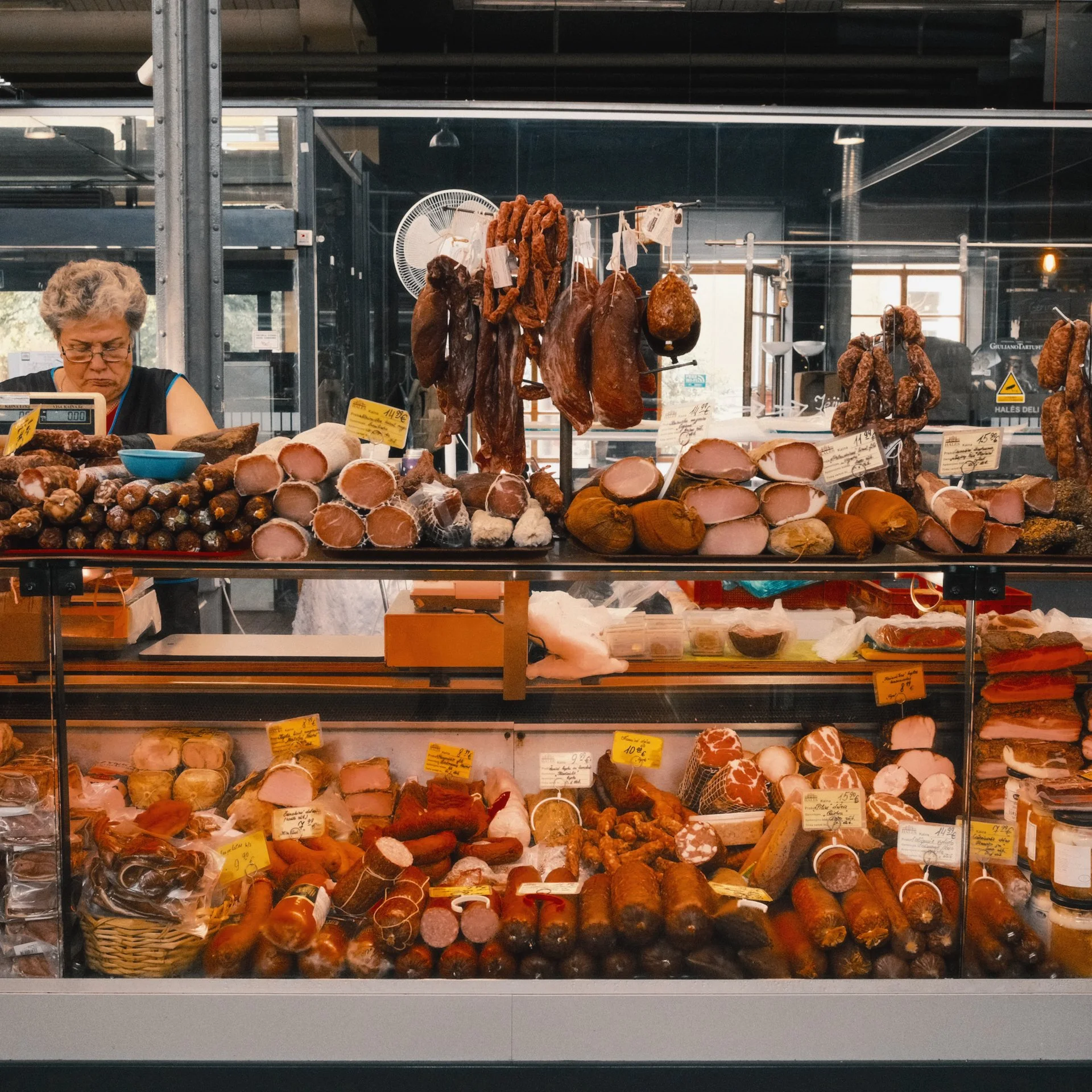 Display of various cured meats and sausages in a butcher shop, with a woman behind the counter and hanging meats and cuts.