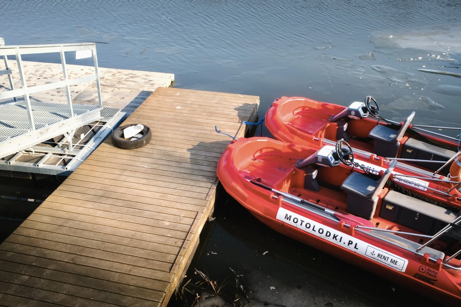 A wooden dock extending into a body of water with two bright orange paddle boats partially tied up at the edge. There is a black tire and a white metal platform on the dock.