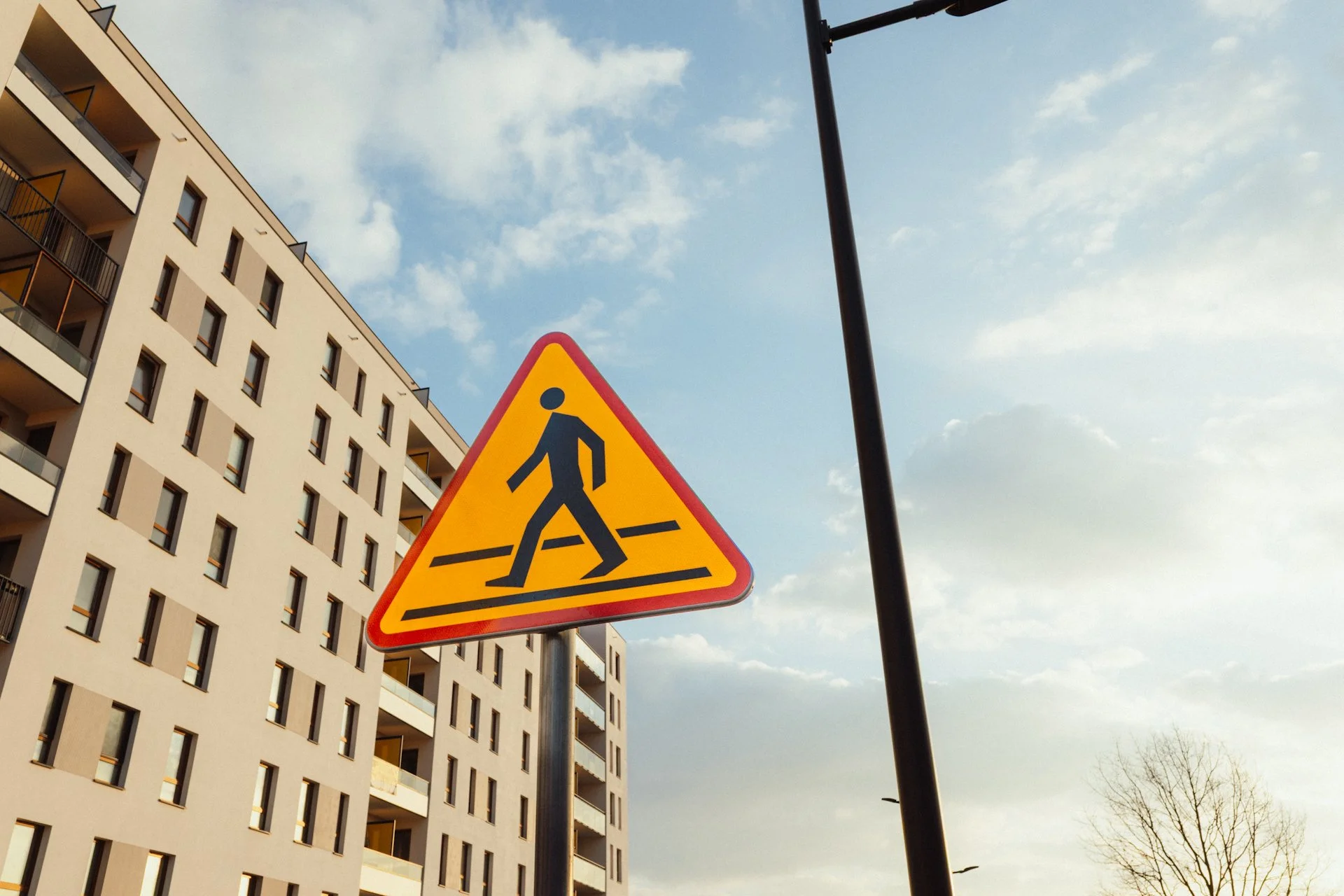 A pedestrian crossing sign on a pole in front of a modern apartment building under a partly cloudy sky.