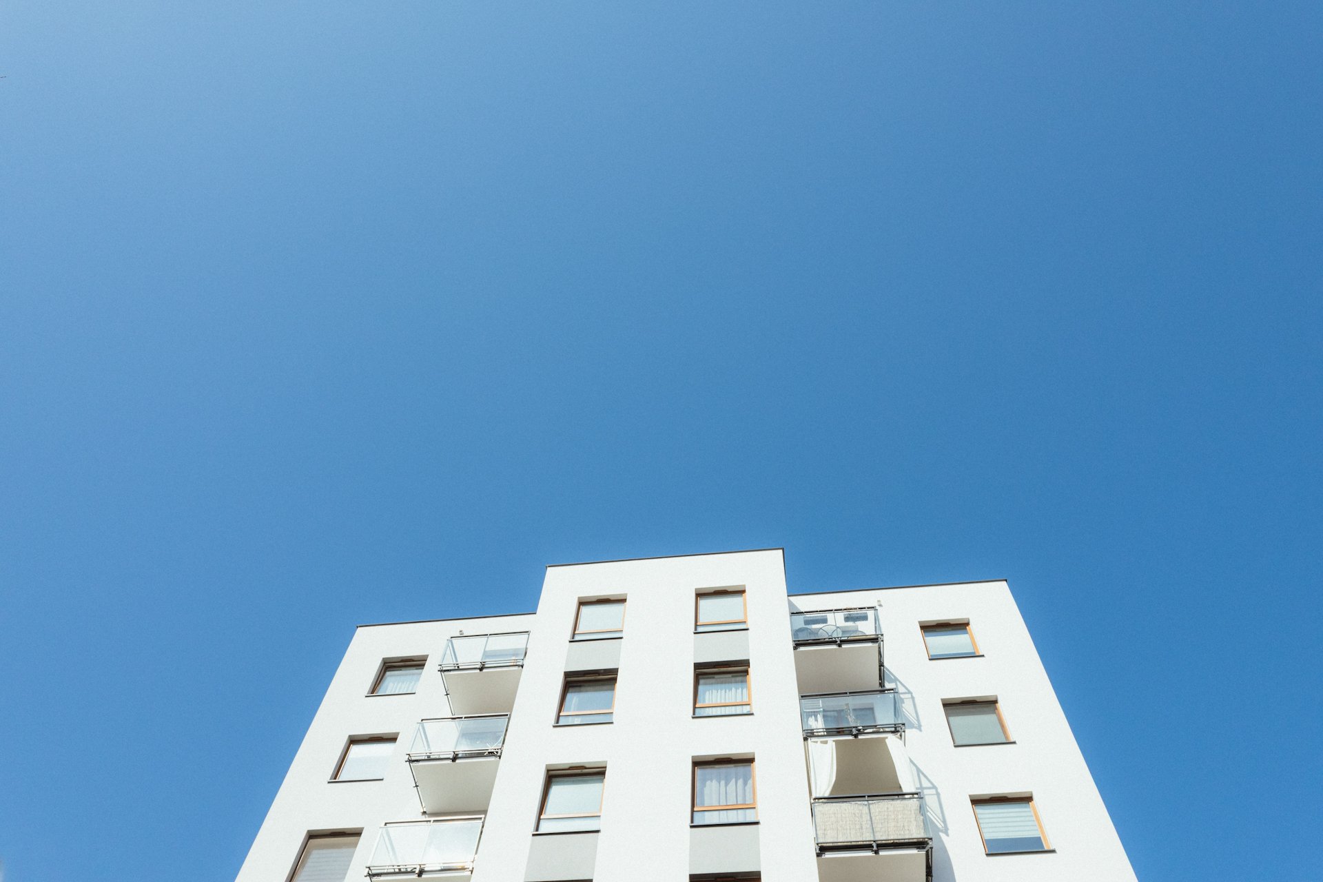 Low-angle view of a modern, white apartment building with several balconies and windows against a clear blue sky.