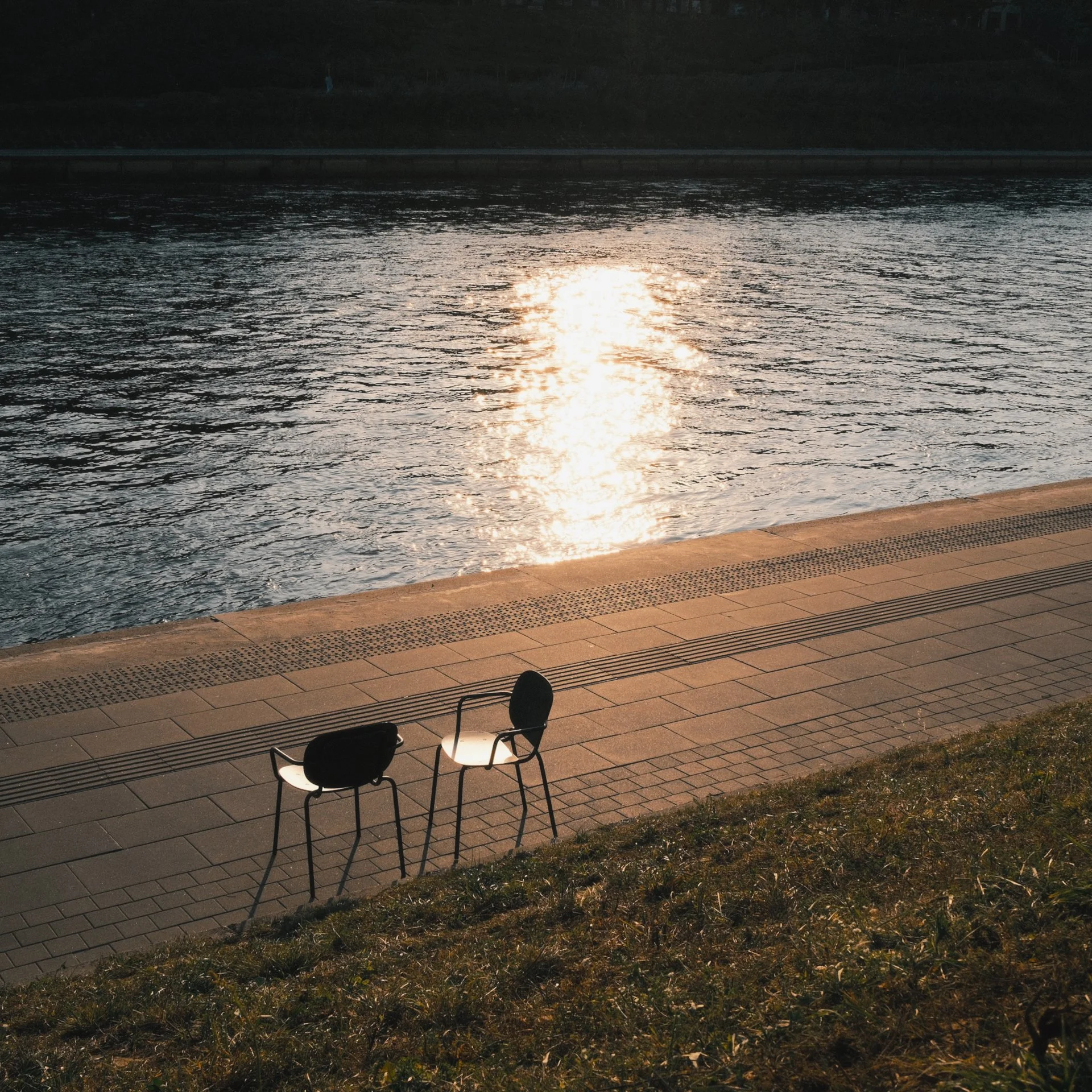 Two chairs facing a water body with the reflection of the sun, located on a paved area next to a grassy patch.