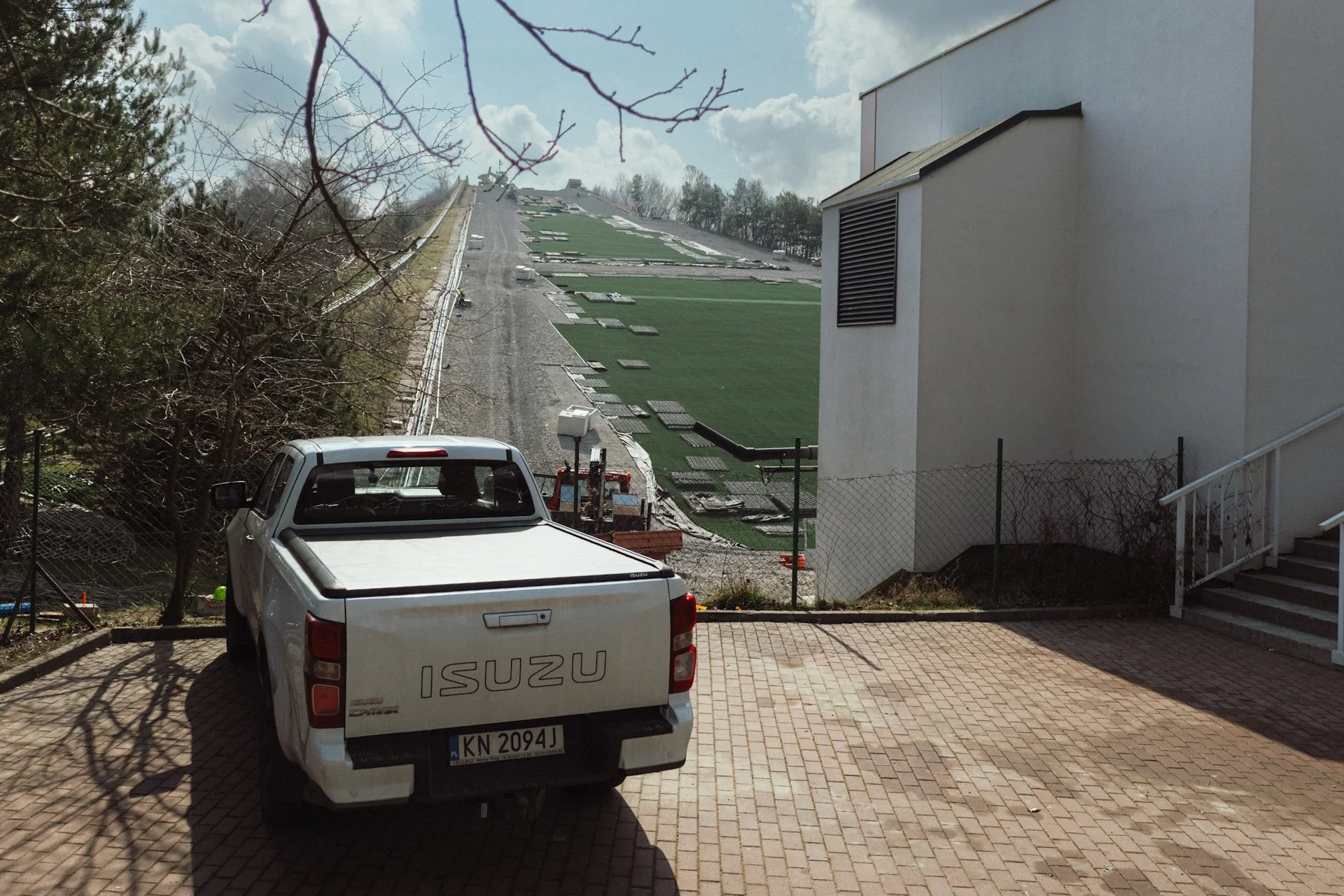 A white Isuzu pickup truck parked in a driveway with a garage building on the right, a chain-link fence, trees on the left, and a steep hillside with construction materials and green artificial turf on the slope in the background.