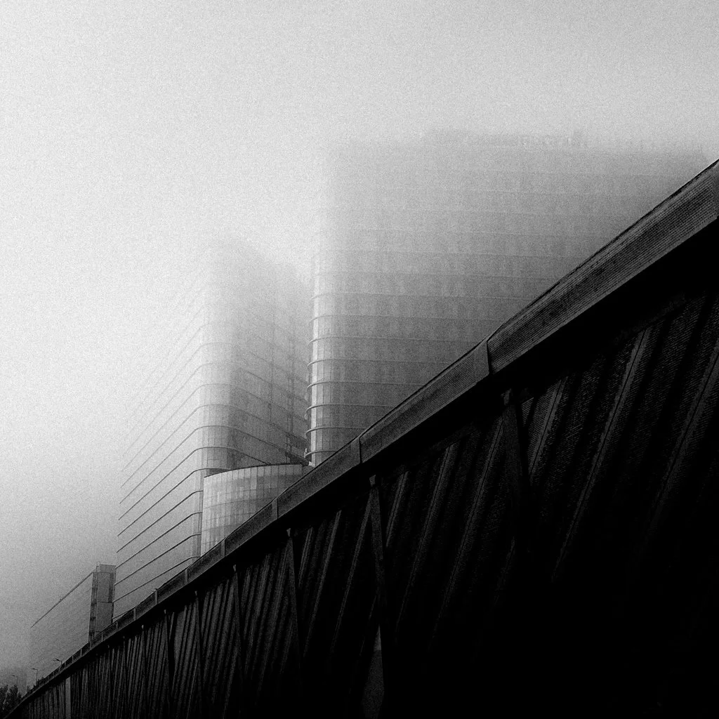 A black and white photo of a modern high-rise building partially obscured by fog, with a bridge or elevated structure in the foreground.