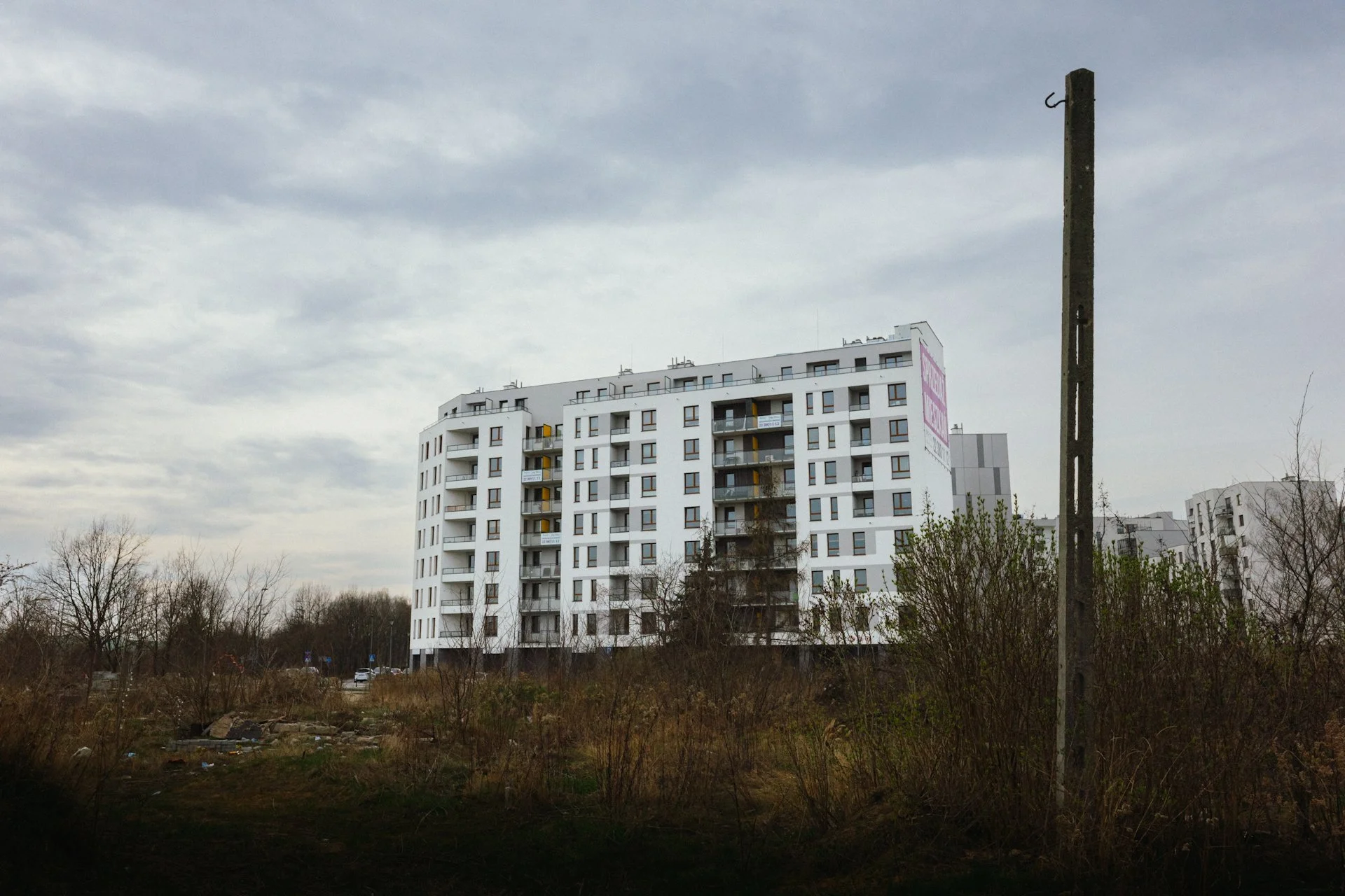 A modern white multi-story apartment building with balconies, situated behind a grassy area with sparse trees and bushes, under an overcast sky.