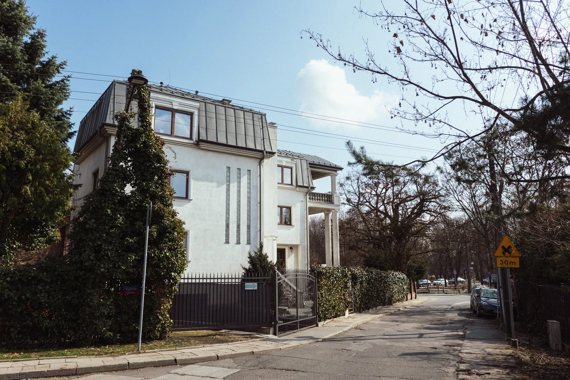 Side street view of a white multi-story house with columns, surrounded by trees and bushes, with parked cars on the street and a yellow traffic sign.