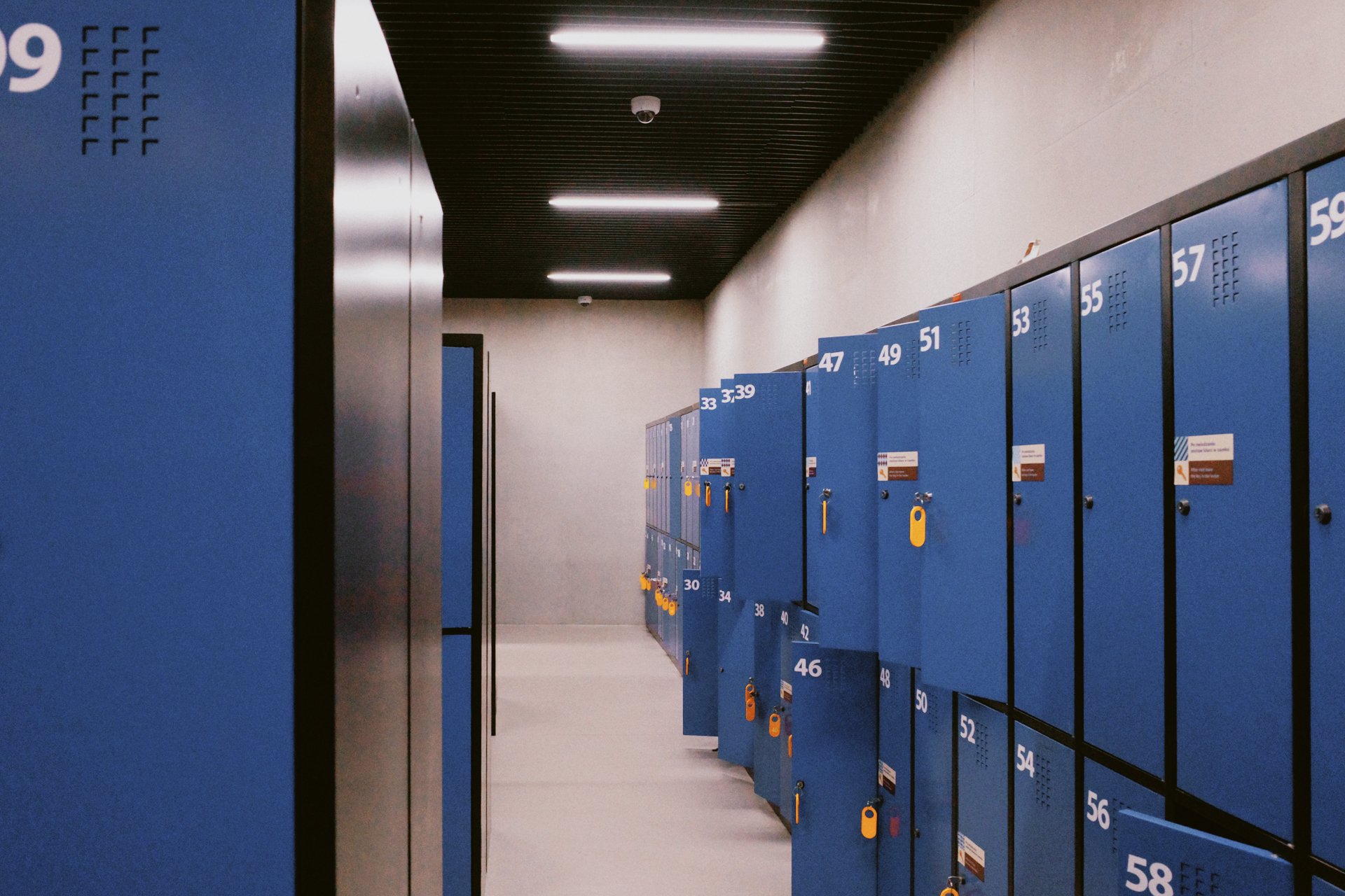 A row of blue lockers in a hallway, numbered from 33 to 58, with some locks secured by padlocks. The hallway has a black ceiling with fluorescent lights and plain white walls.