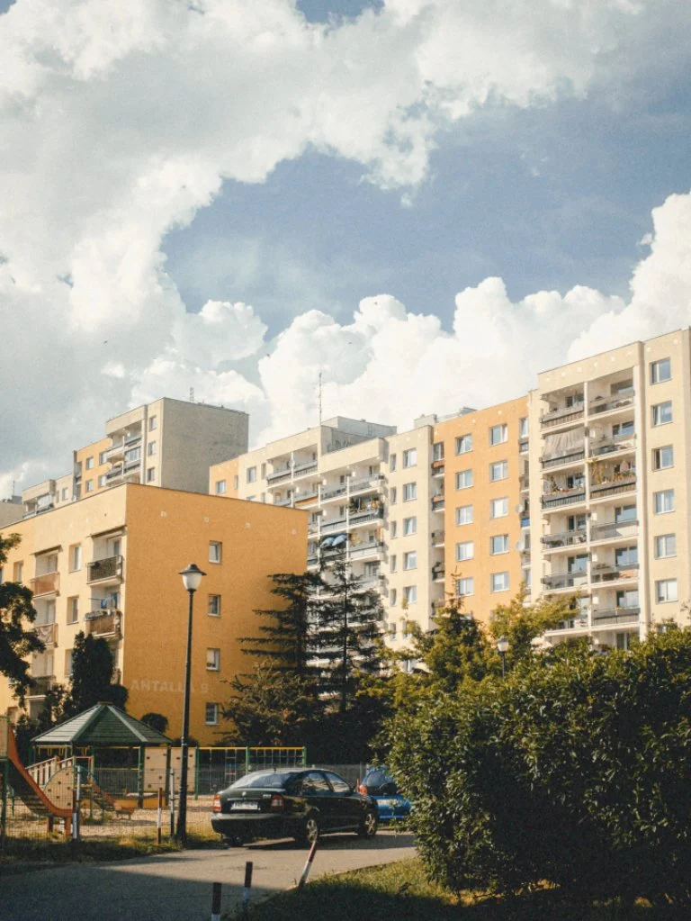 Apartment buildings with balconies, trees, parked cars, a streetlamp, and a small playground under a partly cloudy sky in an urban area.