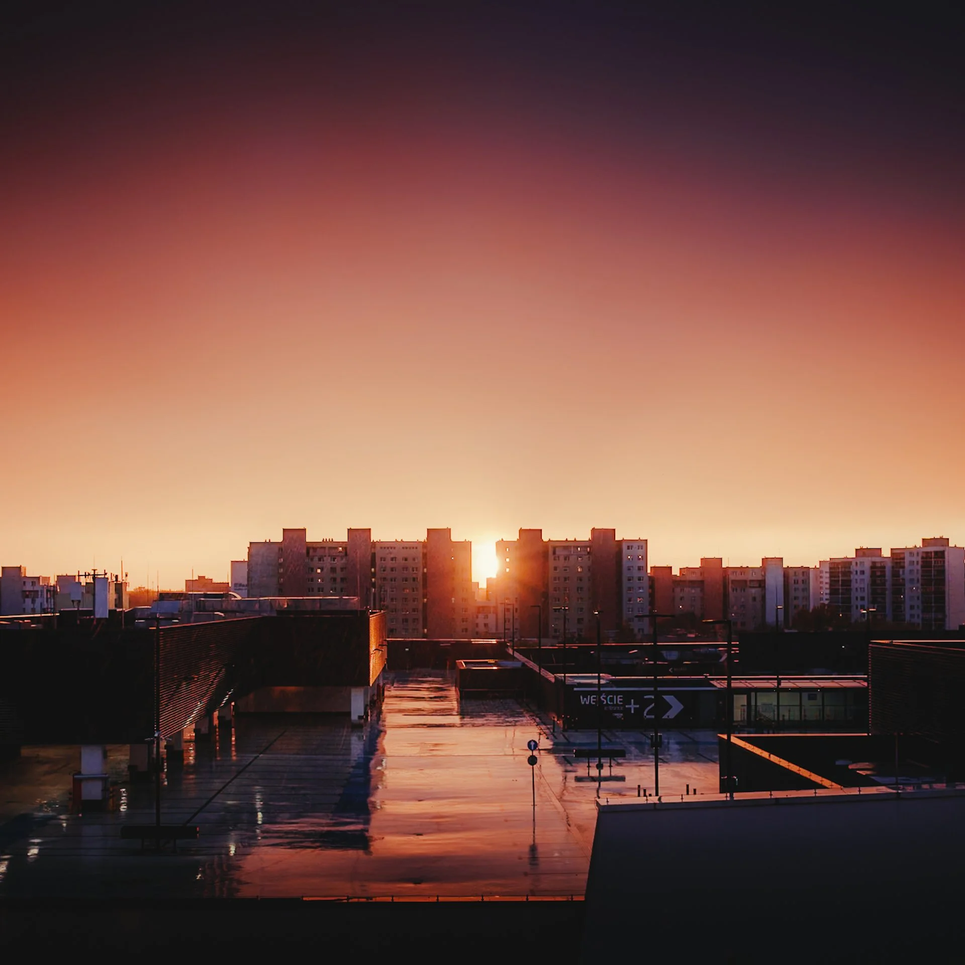 Sunset over a cityscape with high-rise buildings and a wet rooftop reflecting the sky, with a gradient sky transitioning from dark to warm hues.