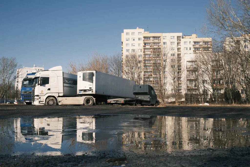 A semi-truck with a white trailer and a black cab at a construction site, with apartment buildings and leafless trees in the background, and a puddle reflecting the scene in the foreground.