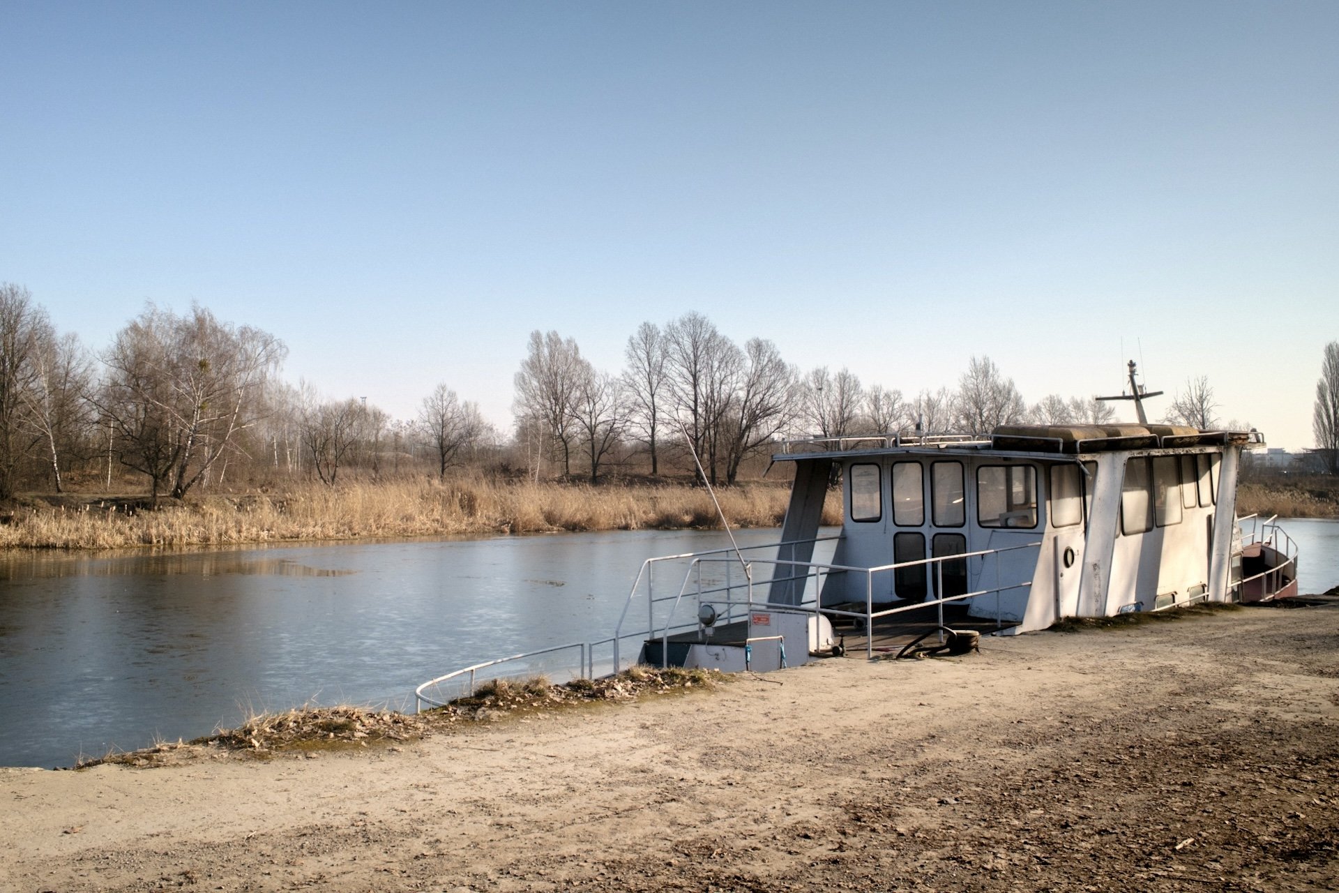 An abandoned boat docked along a waterfront with leafless trees and grass in the background on a clear day.