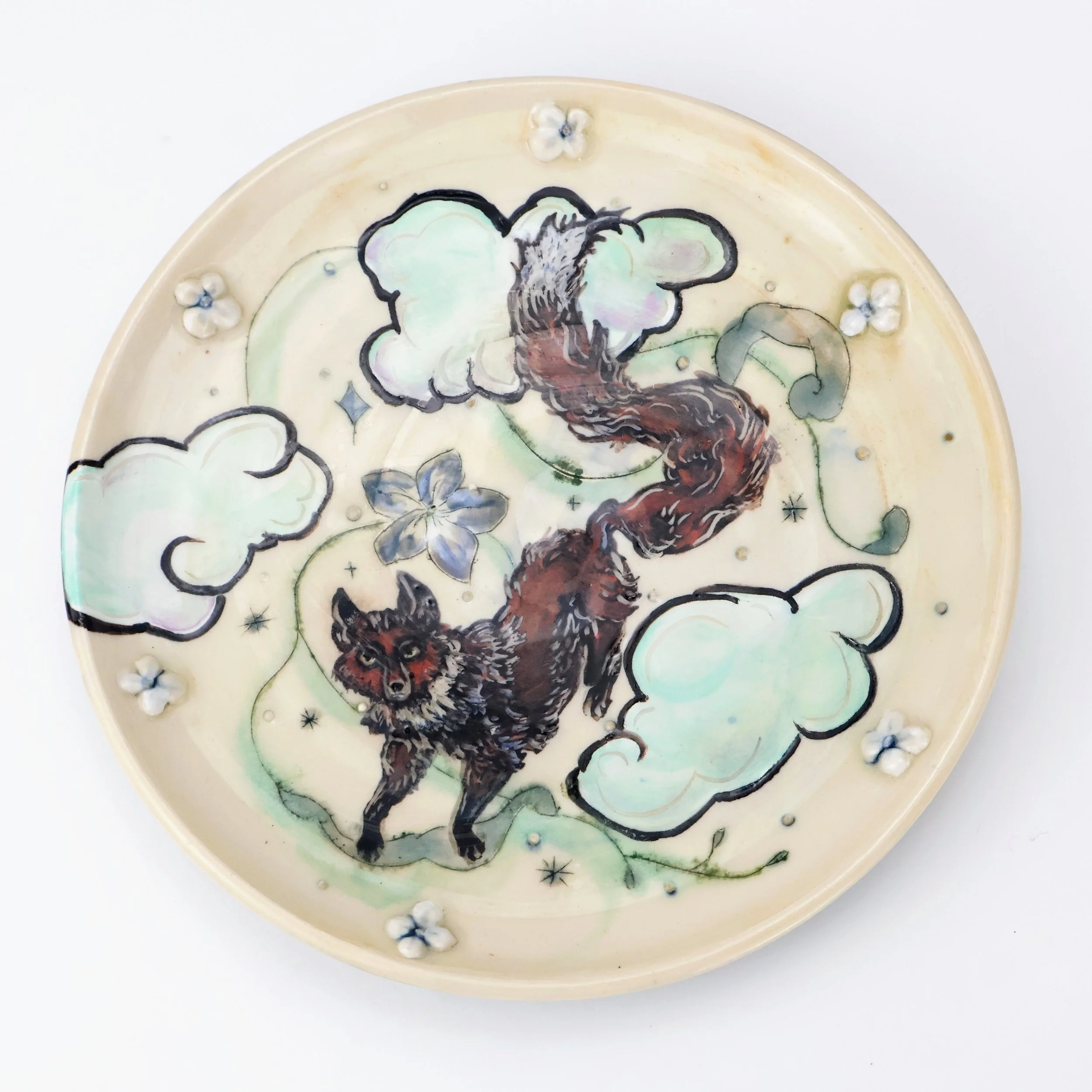 Porcelain plate featuring an illustration of a fox with a long, curling tail, surrounded by clouds, stars, and flowers.