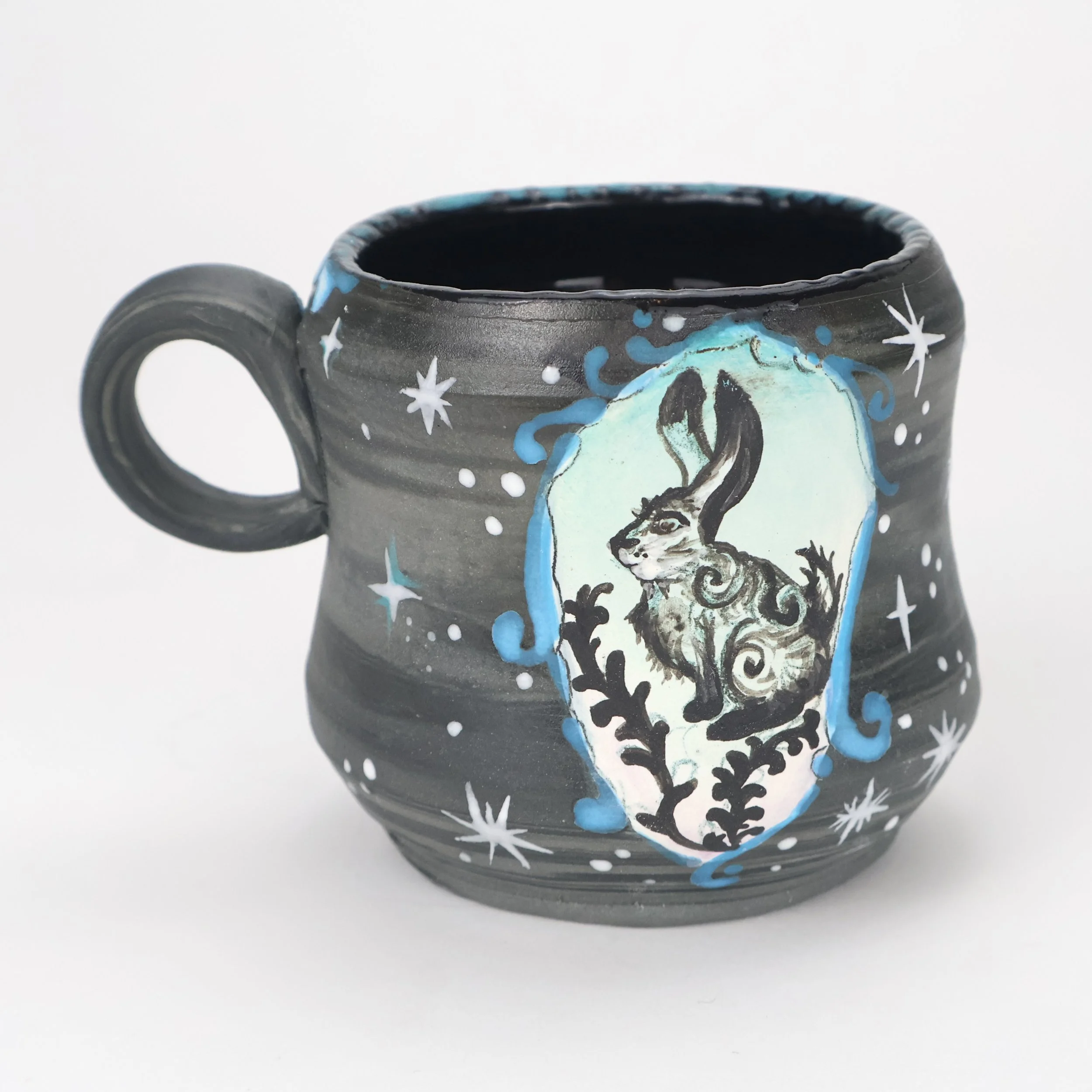 A black ceramic mug with hand-painted magical or mystical design featuring a hare, surrounded by stars and cosmic elements.