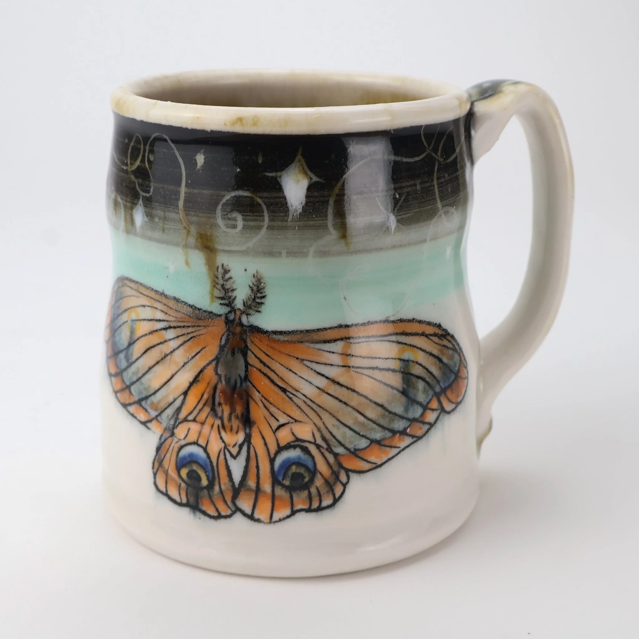 Ceramic porcelain mug with underglaze painting of a large moth with Mishima inlay and a celestial glaze painting, featuring black and blue eye spots on its wings, against a light background.
