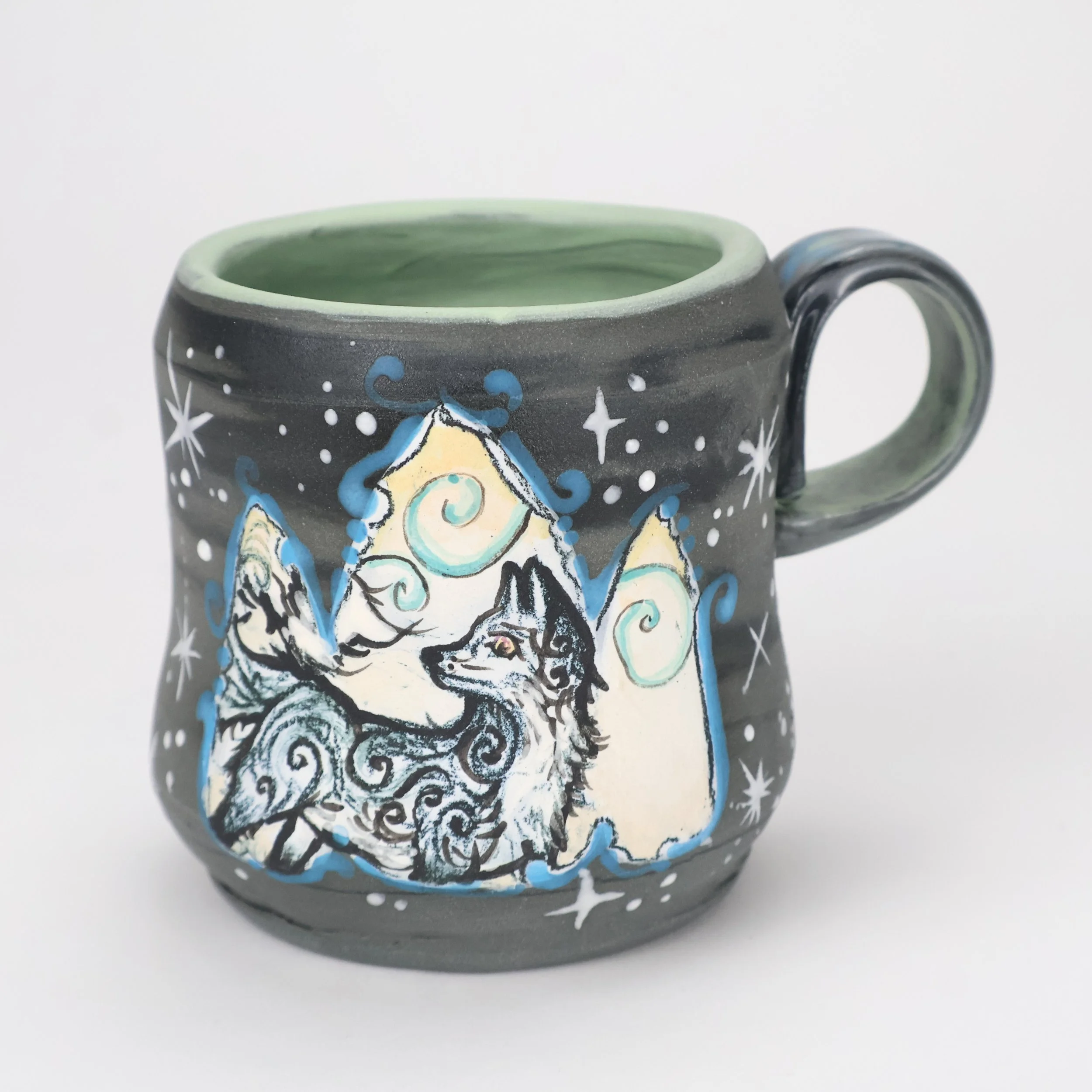 Hand-painted ceramic mug featuring a wolf surrounded by mountains, stars, and swirling clouds with a dark gray exterior and a light green interior.