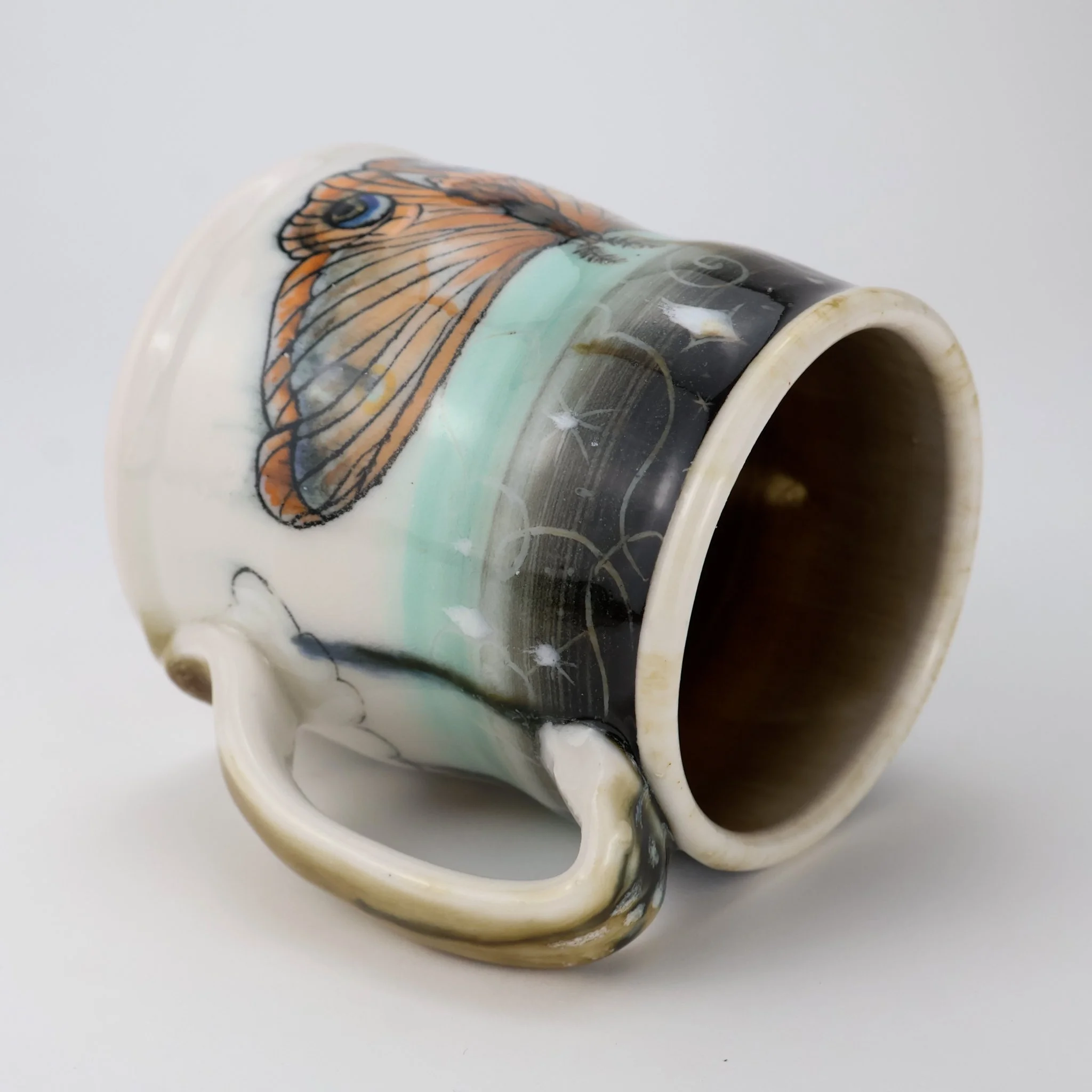 A ceramic porcelain mug with a Moth design and celestial patterns, laying on its side on a plain white background.
