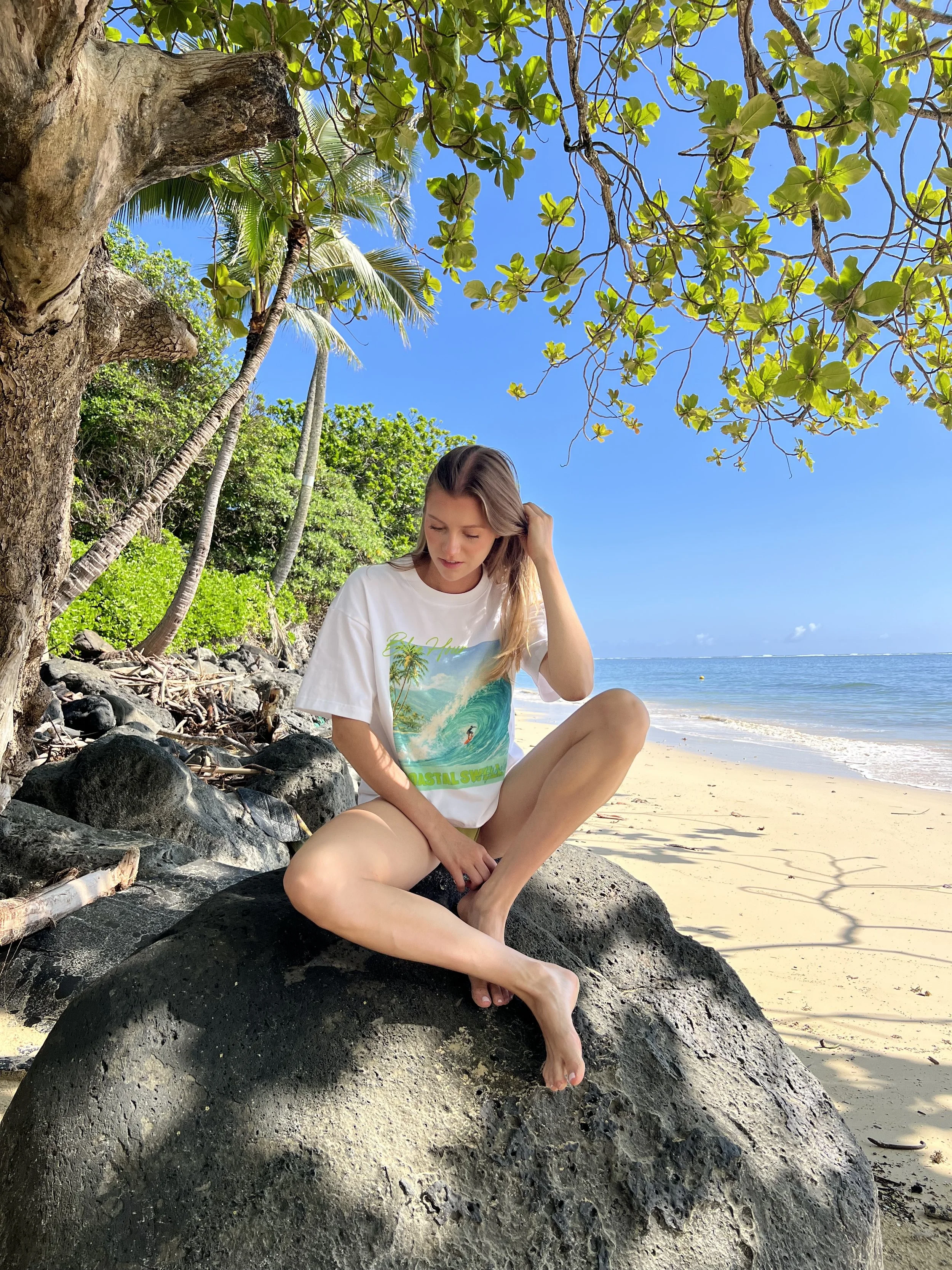 A woman sitting on a large black rock on a beach under a tree with green leaves, with the ocean in the background and a clear blue sky.