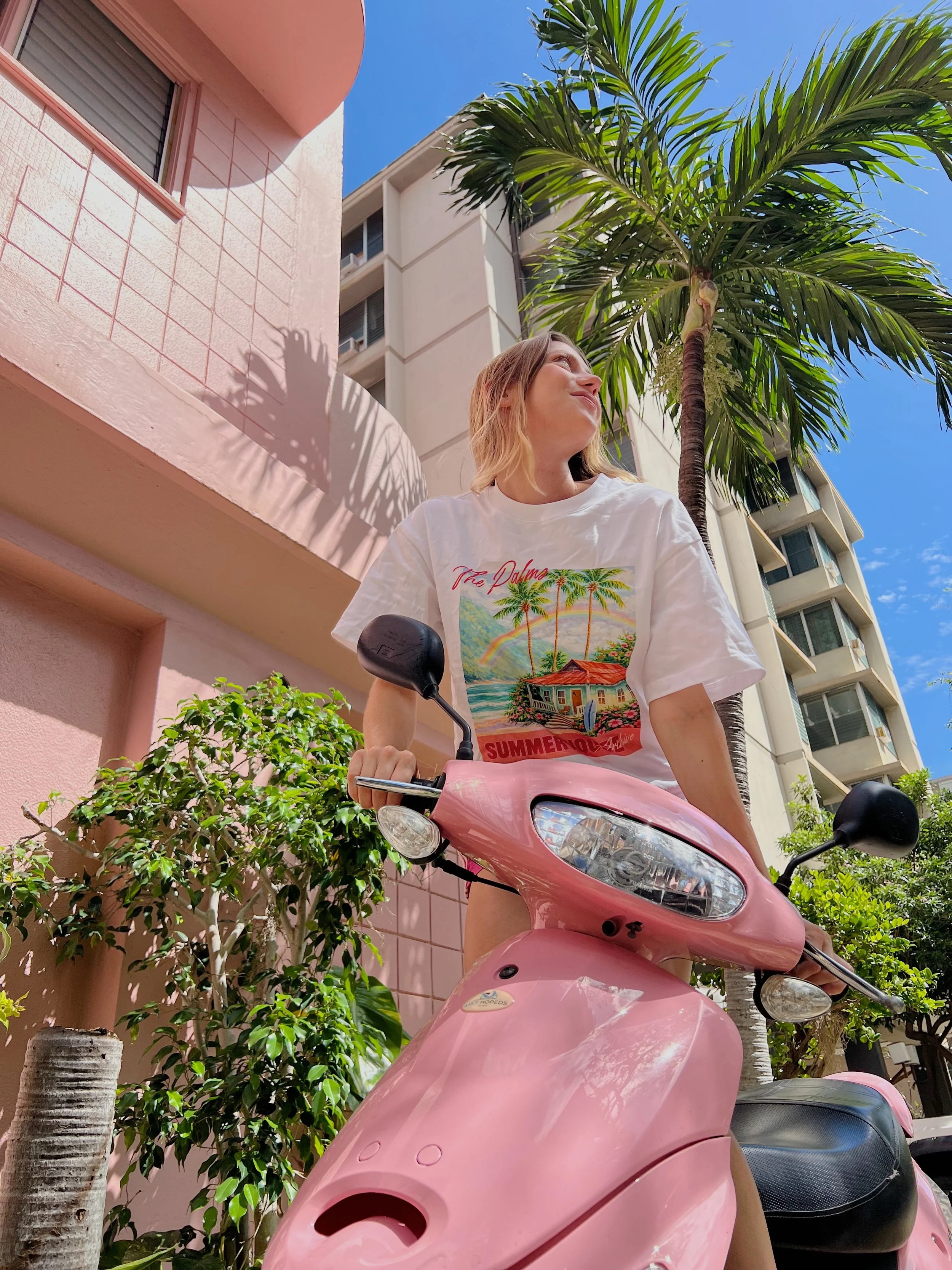 Woman riding a pink scooter outdoors with palm trees and apartment buildings in the background on a sunny day.