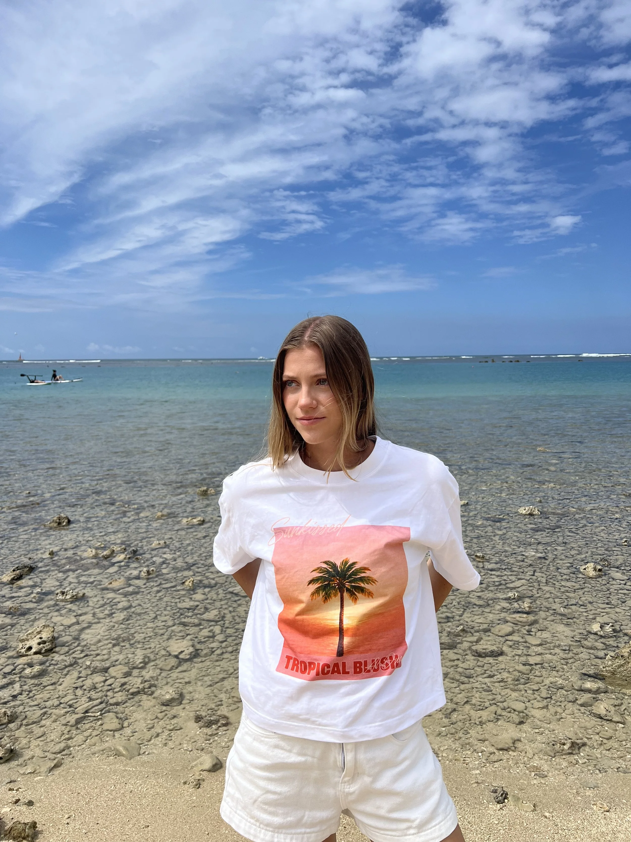 A young woman with blonde hair, wearing a white T-shirt with a palm tree graphic and the words 'Tropical Bliss', standing on a rocky beach with the ocean and blue sky with scattered clouds in the background.
