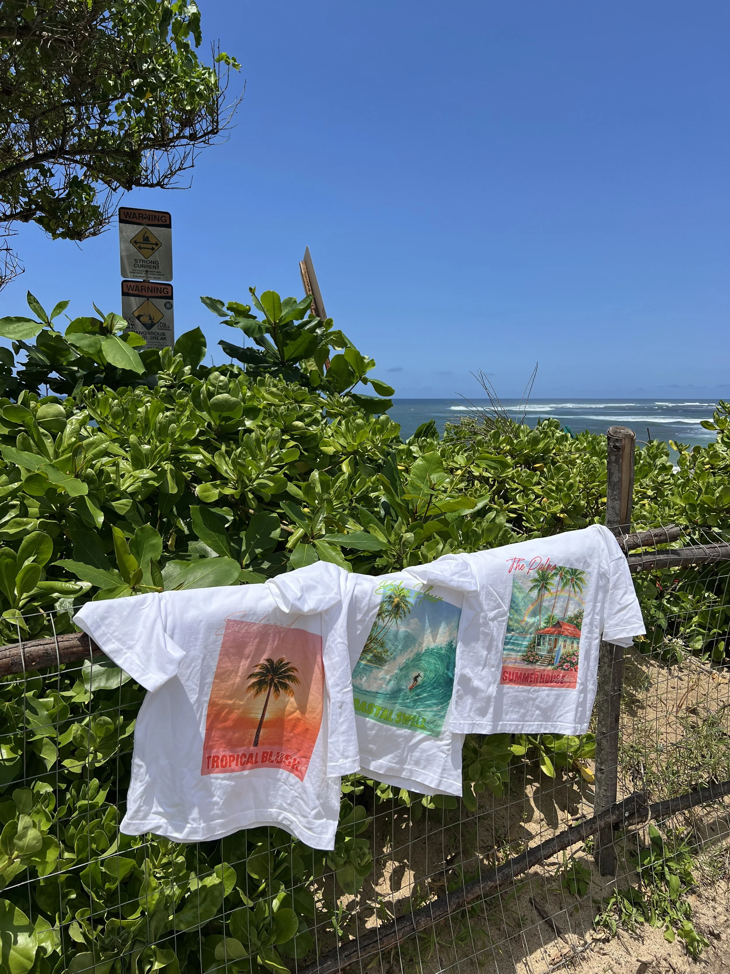 Three white T-shirts with colorful beach-themed prints are hanging on a wire fence at a tropical outdoor location with lush green foliage, a sandy ground, and the ocean in the background under a clear blue sky.