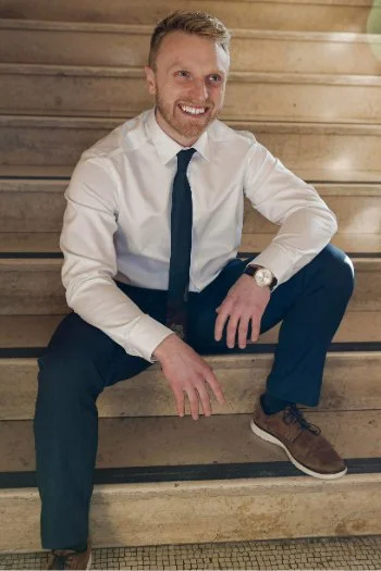 Man in white shirt and dark tie sitting on wooden stairs, smiling, with a watch on his left wrist.