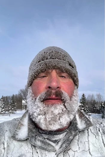 A man with a snow-covered beard and face, wearing a gray knit hat and gray jacket, outdoors in a snowy landscape with trees and a cloudy sky.
