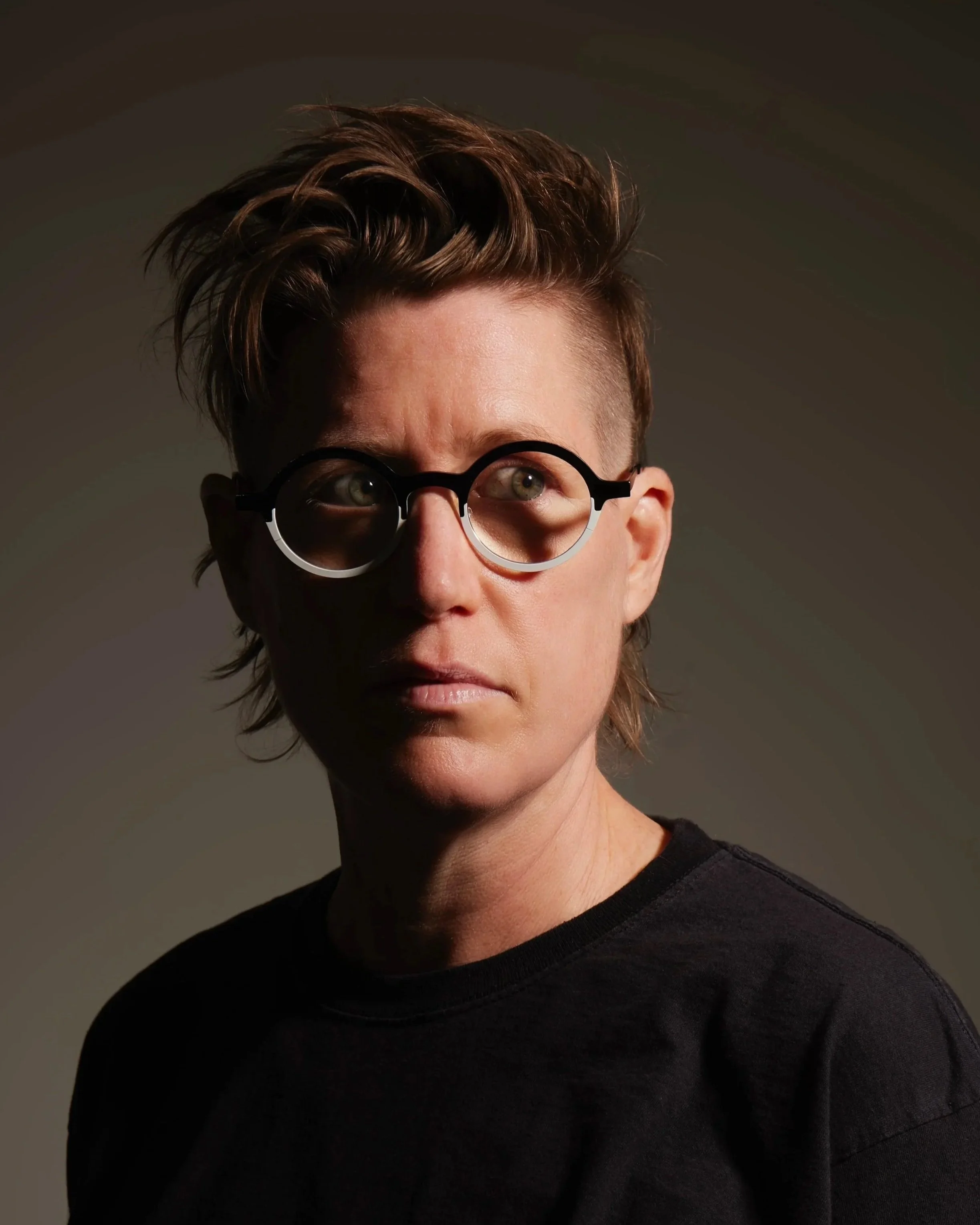 A person with short, wavy hair wearing round glasses and a black shirt, looking slightly to the side with a serious expression, against a gray background.