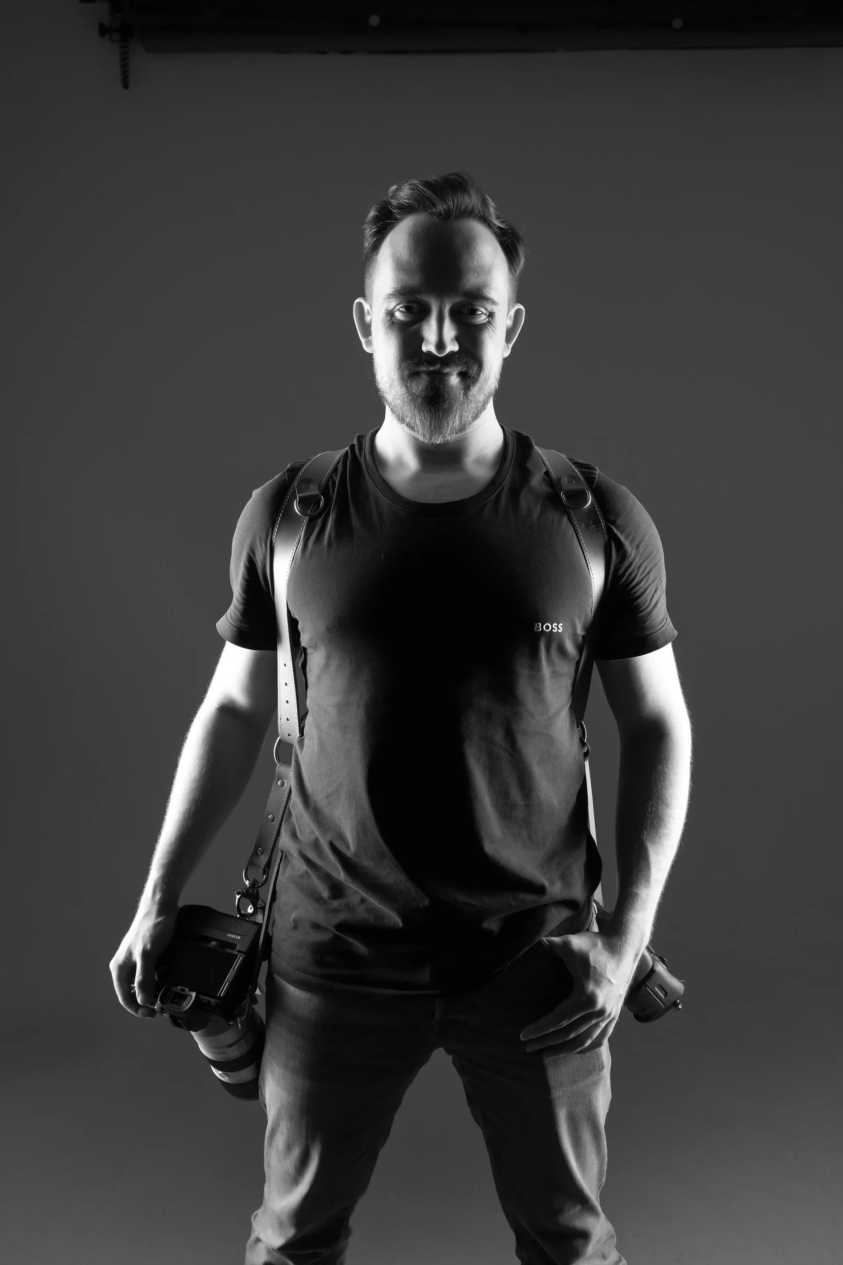 Schwarz-weiß-Fotografie eines jungen Mannes mit einem Rucksack, der eine Kamera in der Hand hält, vor einem dunklen Hintergrund.