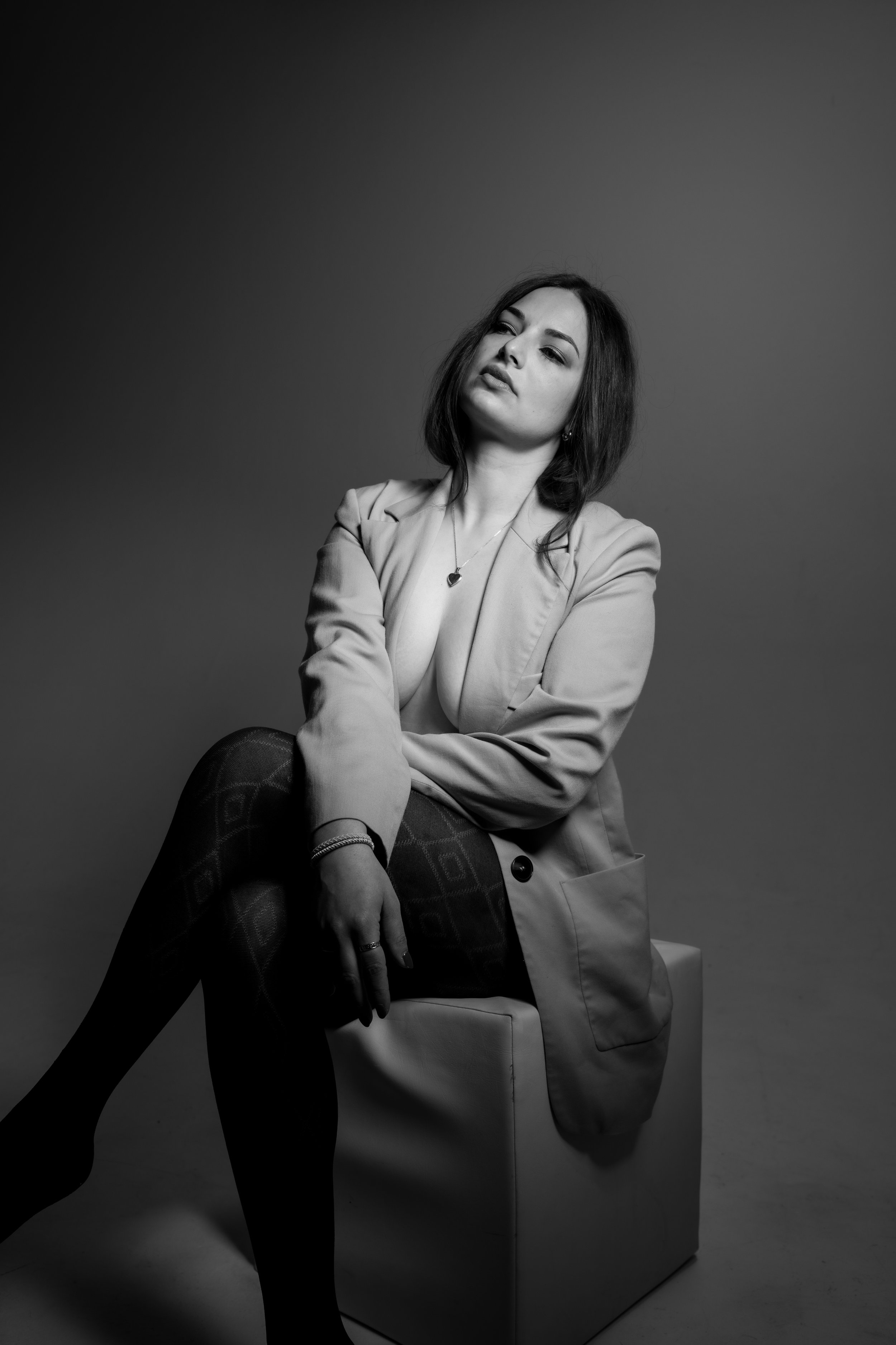 Schwarzweiß-Boudoirfoto einer Frau im Blazer und Strumpfhosen vor grauem Hintergrund