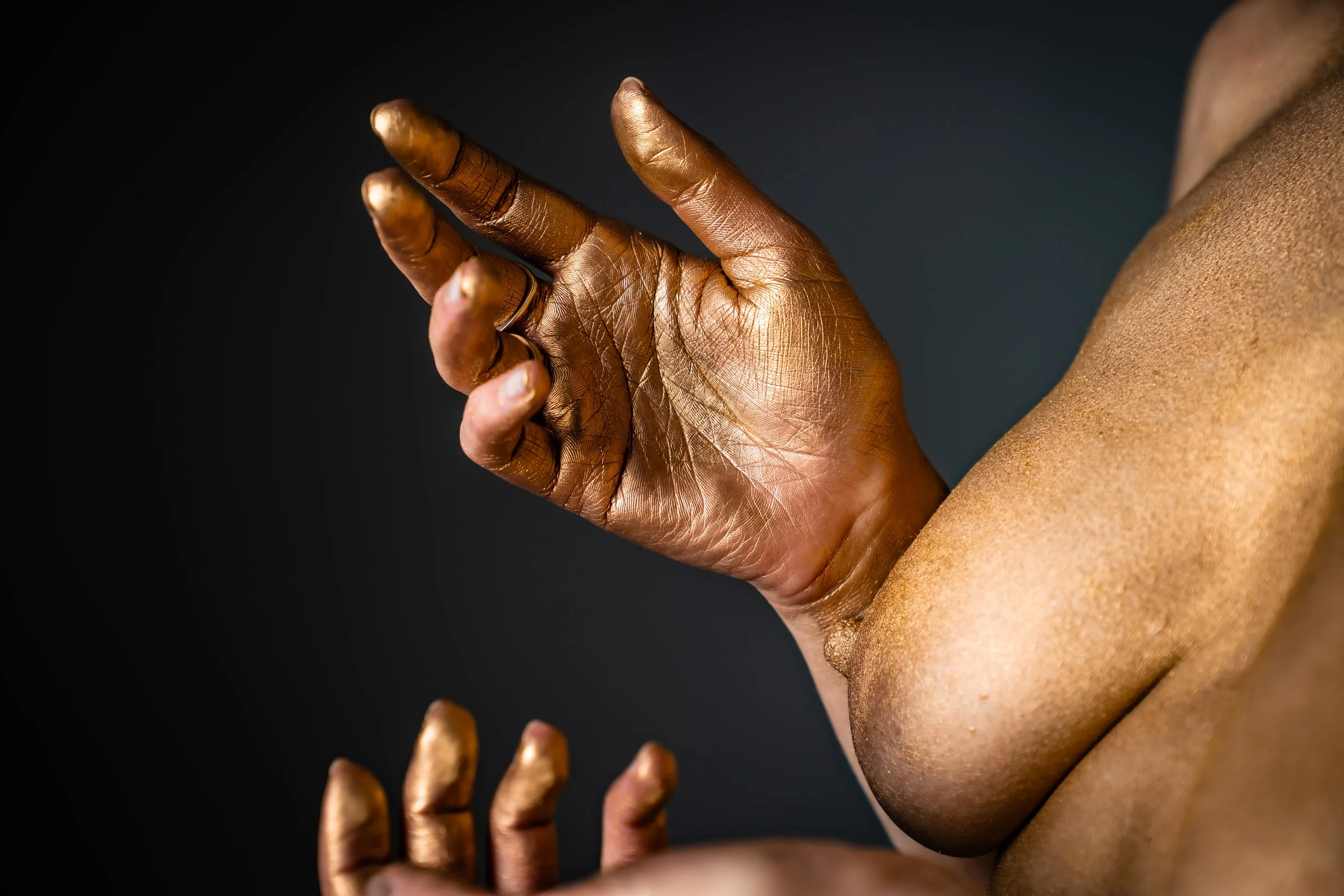 Nahaufnahme einer menschlichen Hand mit vollständig lackierter goldener Haut, die in einer Geste gehalten wird, vor einem dunklen Hintergrund.