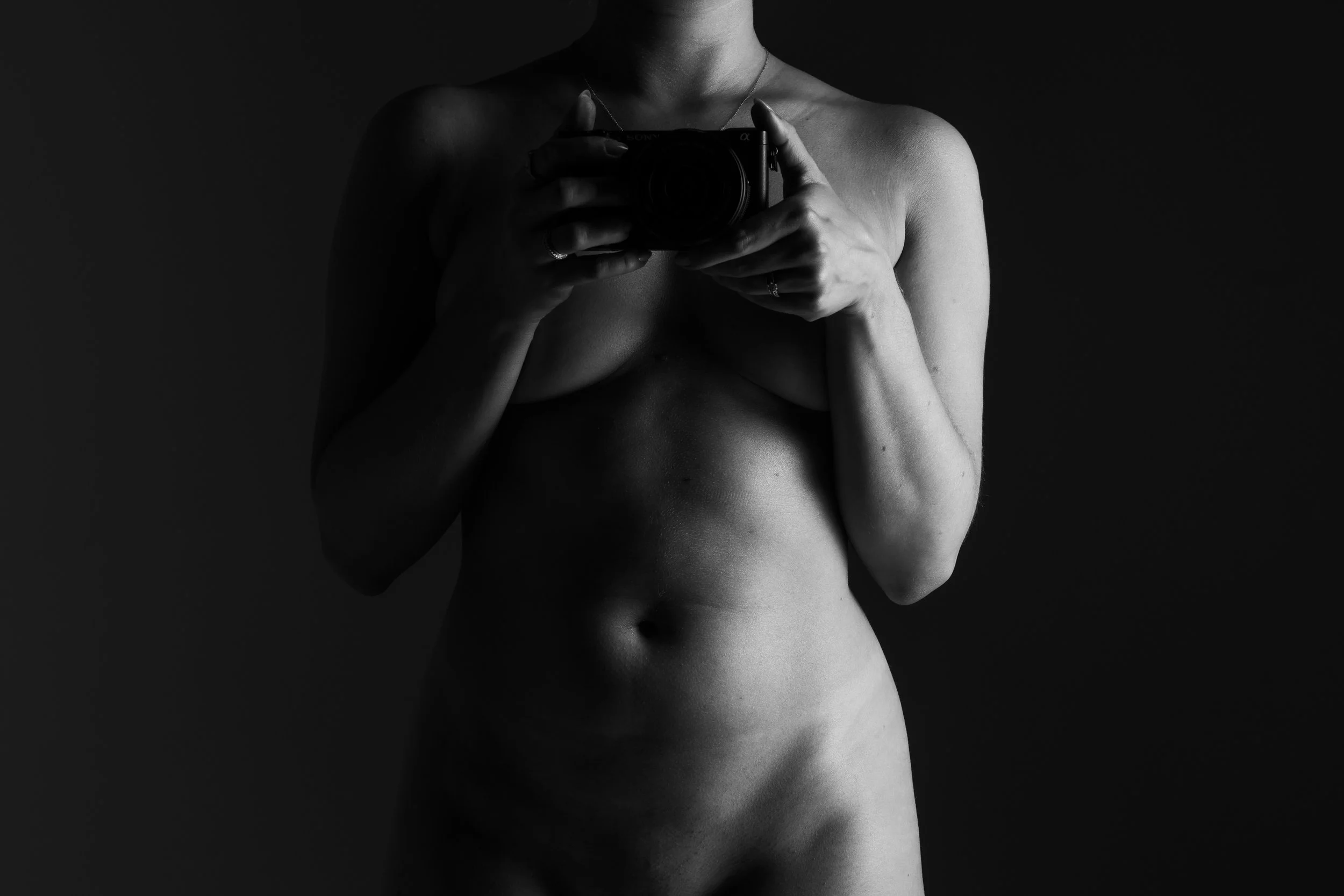 Schwarz-weiß- Aktfotografie eines Frauenkörpers, aufgenommen im Selbstporträtstil, mit Fokussierung auf den Oberkörper und die Mitte des Bauches, gehalten vor dunklem Hintergrund.