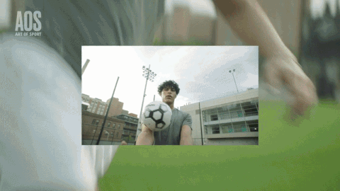 A soccer player on a field with a stadium and city buildings in the background, preparing to kick a soccer ball.
