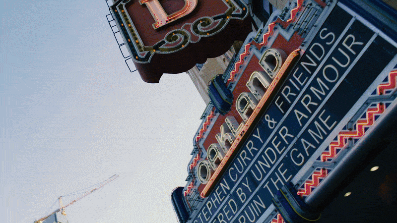 Theatrical marquee signs for the Broadway musical "Chicago" with additional signage about curry and friends under armor, featuring colorful neon lights and a cloudy sky.