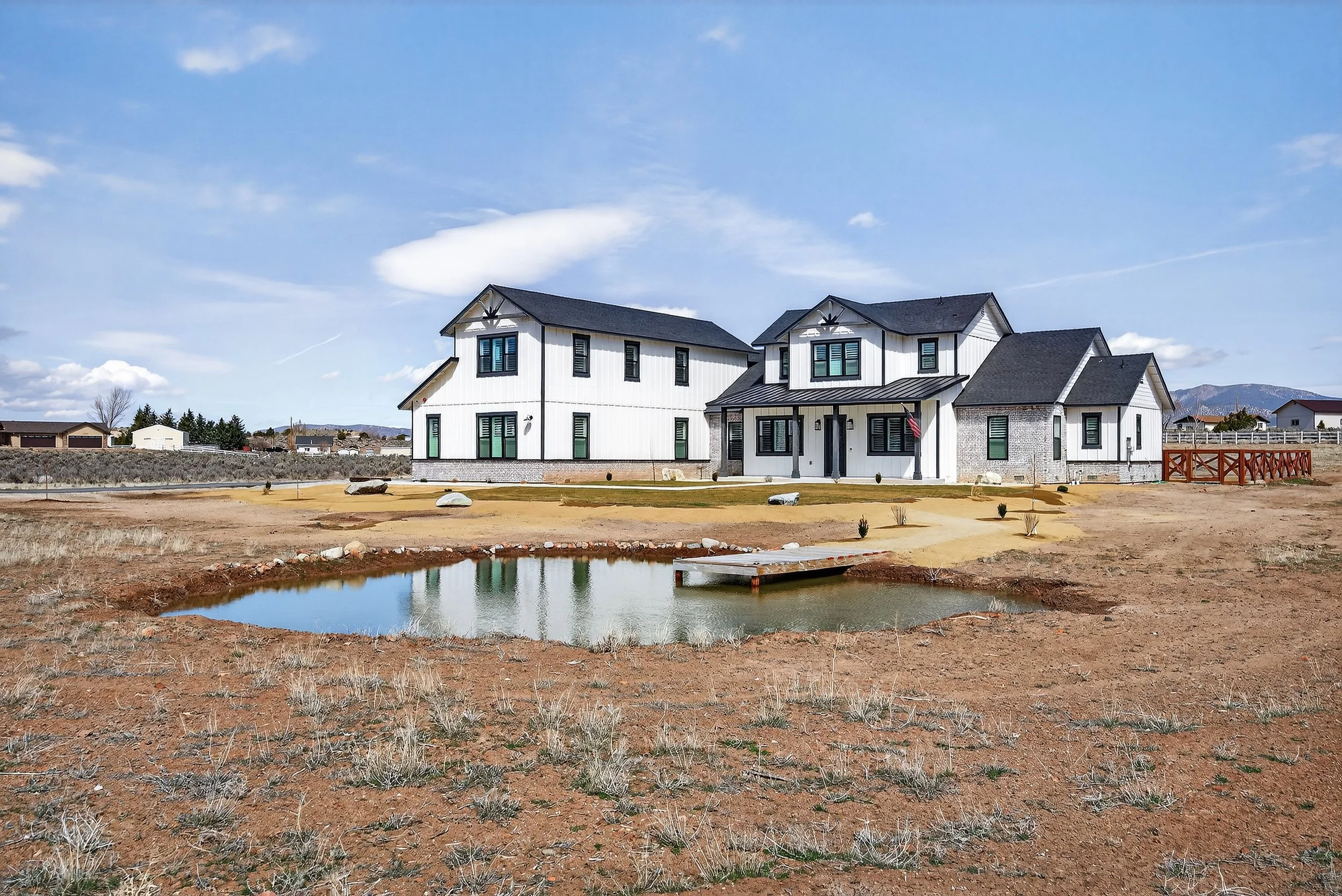 A modern two-story house with white vertical siding, black window shutters, and a gabled roof, situated in a rural area with dry grass, a small pond, and mountains in the background.  Carson Valley, Minden, Gardnerville, Nevada.