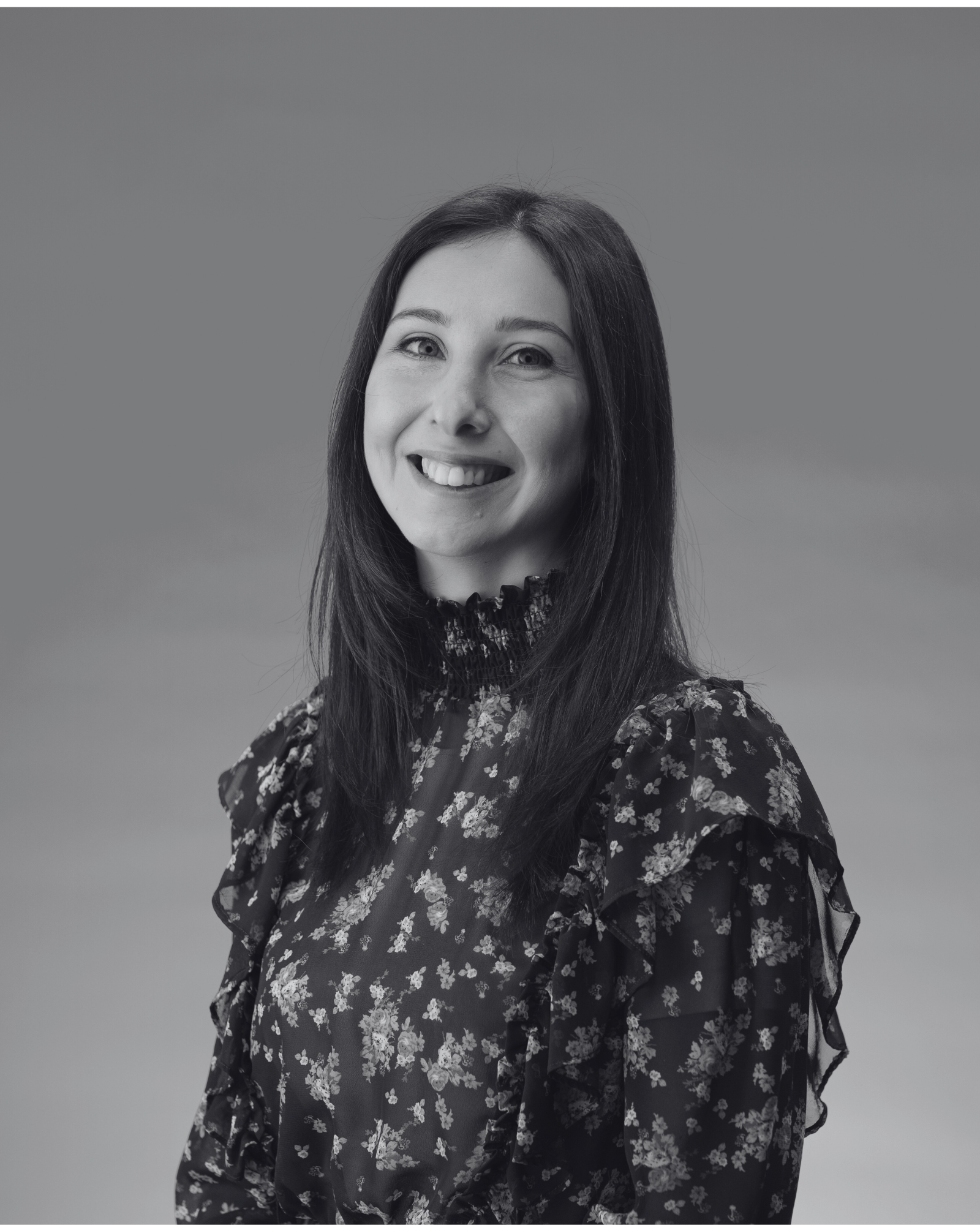A black and white portrait of a smiling woman with long dark hair wearing a floral blouse against a plain background.